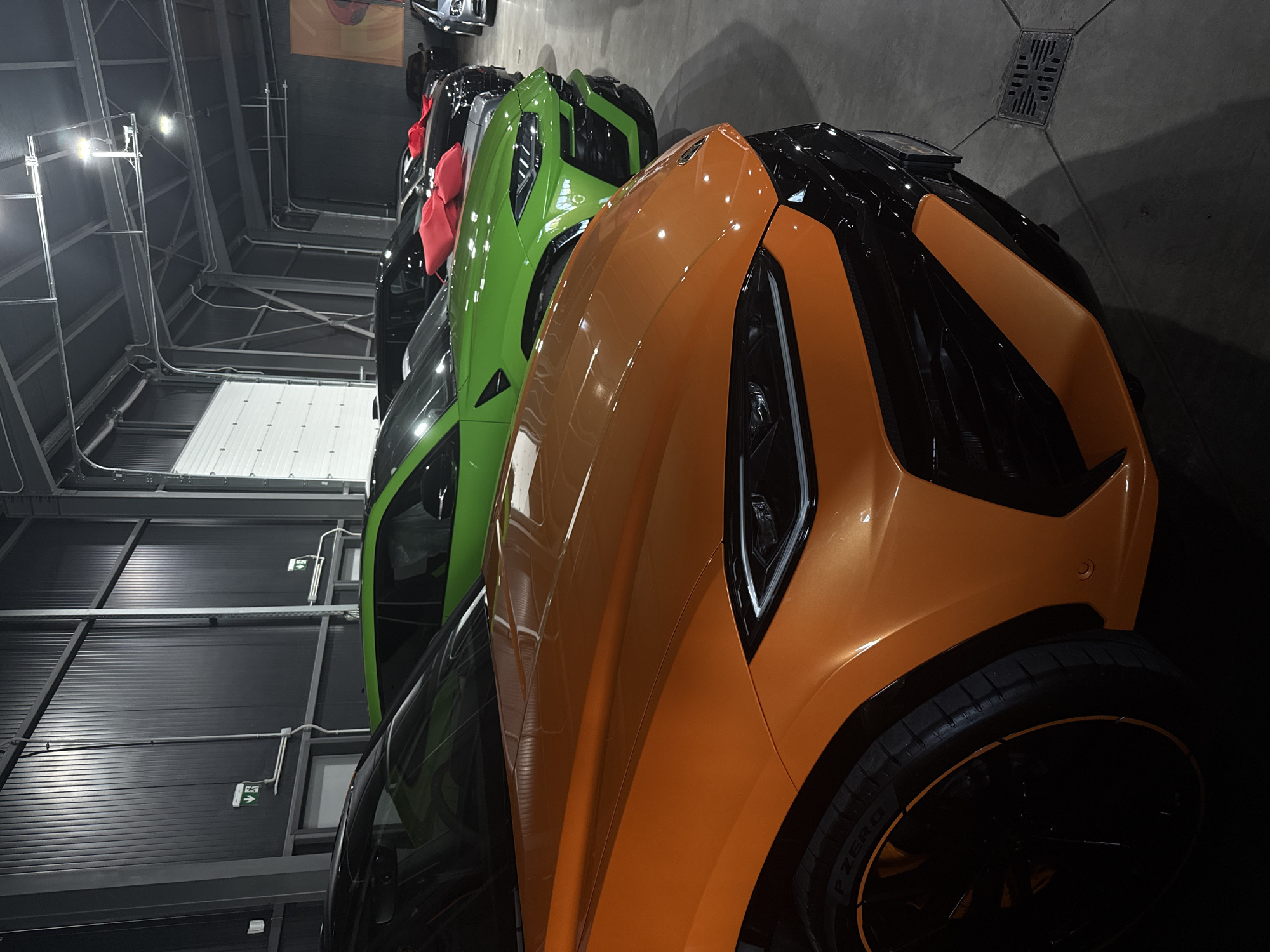 The image shows a lineup of luxury vehicles, primarily Lamborghini Urus models, parked indoors in what appears to be a garage or showroom. The colors of the vehicles include orange, green, and black. The setting is likely an underground parking garage or enclosed space in Plovdiv, Bulgaria, although specific details confirming Plovdiv are absent. The scene features a car showroom or garage setting with industrial lighting. There are no people visible.