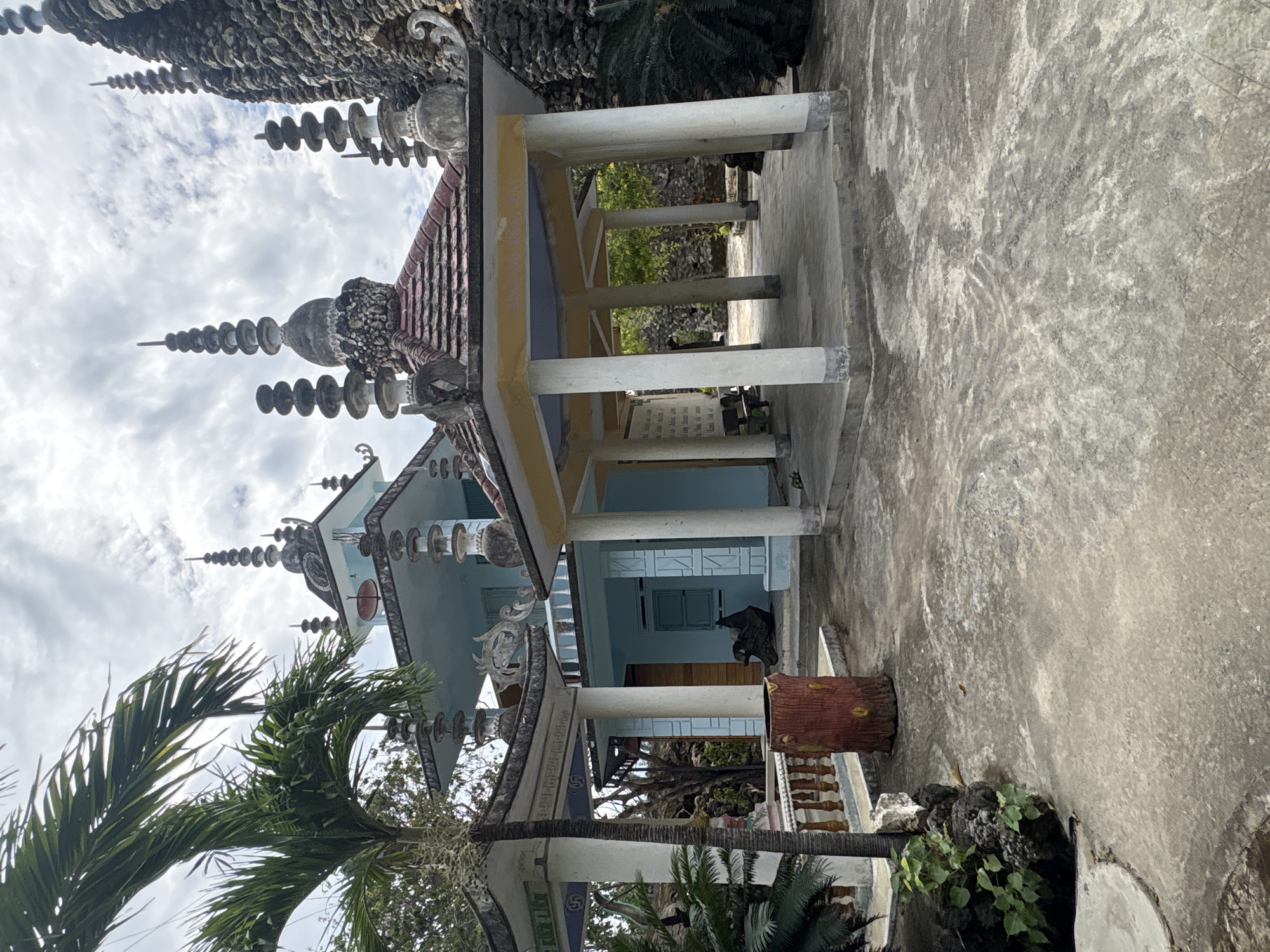 This image captures an ornate architectural structure, likely a temple or pagoda, under a cloudy daytime sky in Cam Ranh, Vietnam. The building features a distinctive light blue facade and a prominent dark, textured roof adorned with multiple elaborate spires and decorative finials. These roof decorations consist of stacked, disc-like elements that taper upwards, alongside intricate, possibly shell-encrusted, sculptural details.
A large, open porch or veranda, supported by several light-colored pillars, extends from the main building, showcasing a light yellow underside. Below this porch, light blue doors and windows are visible, and a dark, draped object rests near one of the doorways. On the ground level, a broad expanse of rough concrete or paved surface stretches across the foreground and to the right. A reddish-brown, log-shaped planter is situated on the right side of the frame.
Lush tropical vegetation, including large palm trees with their green fronds, frames the building from the left and lower portions of the image. Other green foliage and dark, rocky formations are scattered around the building's base. The sky above is overcast or partly cloudy, indicating daylight. A white sign with black text, likely Vietnamese, is mounted on a wall under the porch, though its content is illegible. No people or specific activities are depicted, focusing instead on the unique architectural details and natural surroundings of this cultural site.