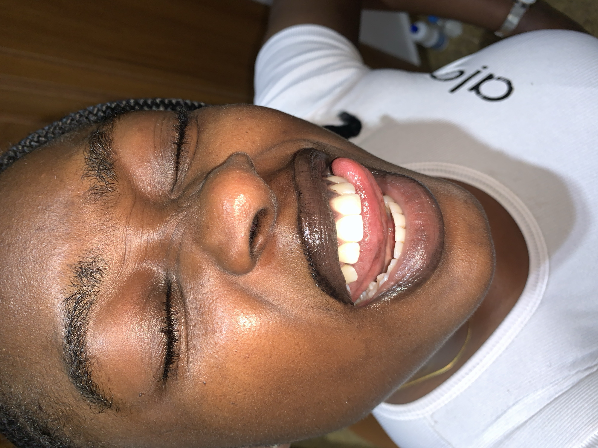 A close-up, indoor, waist-up shot captures a young Black woman with braided hair wearing a white t-shirt and a gold necklace. Her eyes are closed, and her mouth is open, with her tongue extended and visible. Her lips are glossed with a dark lipstick, and her teeth are white. The background is blurred but appears to be a room with wooden paneling and some objects on a surface. The lighting is warm and appears to be from a flash. There is no indication of the location being Gura Top, Nigeria.