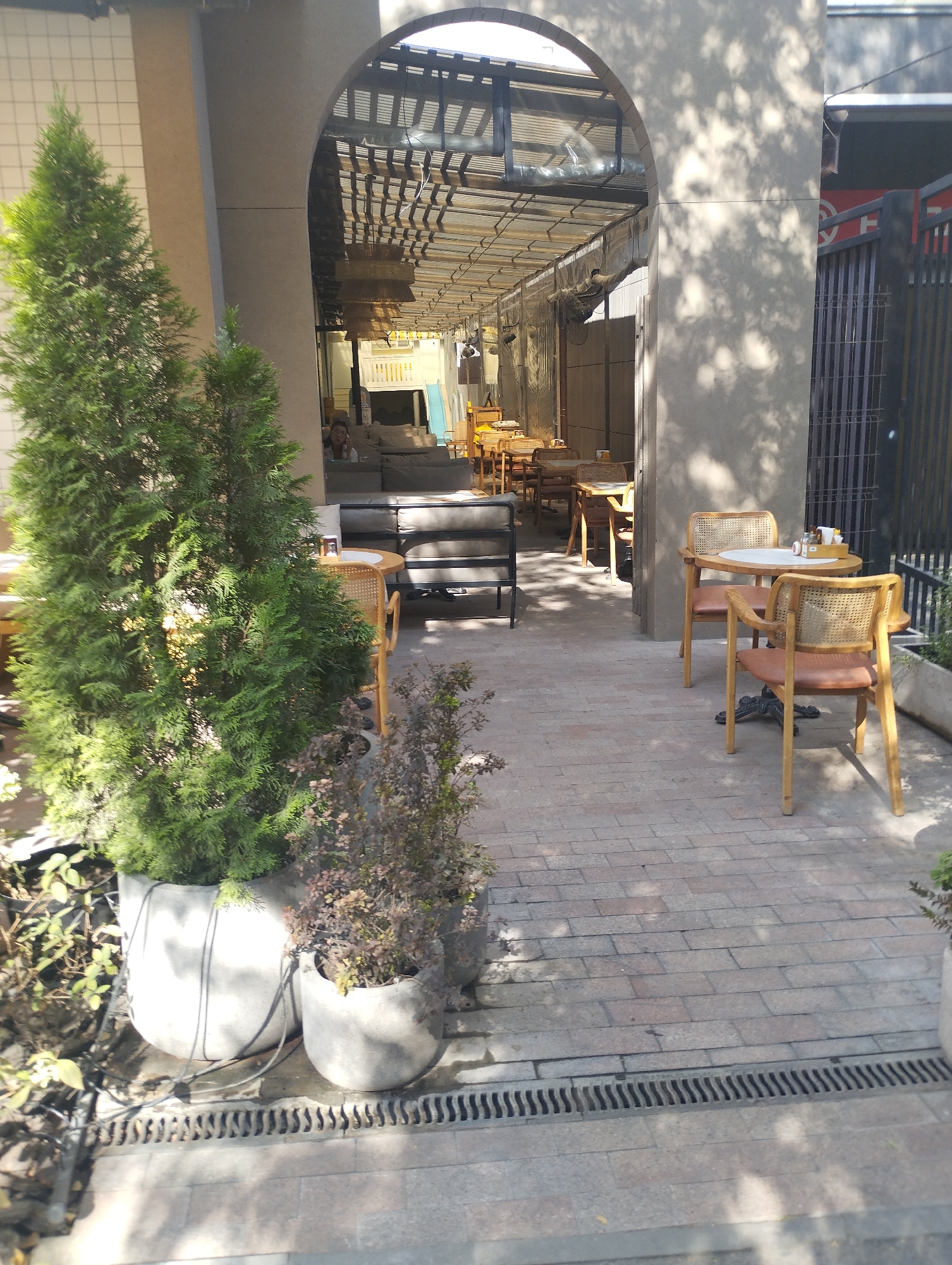 This is an eye-level, medium shot of an outdoor seating area of a cafe or restaurant. The scene is set in Bishkek, Kyrgyzstan. In the foreground, two large potted evergreen trees flank a paved walkway made of brick-like stones. A black metal grate runs along the edge of the walkway. To the right, a small round table is set with a salt and pepper shaker and a small rectangular container, accompanied by two cane-backed chairs with light brown leather seats. Further back, through an arched entryway, is another seating area with more tables and chairs, some of which are wooden and some are upholstered sofas. The ceiling of the indoor/outdoor space is covered with a metal grate structure, and overhead lighting fixtures are visible. Sunlight casts dappled shadows across the patio. A person is visible seated on a sofa in the background. The overall impression is a bright, airy, and casual outdoor dining space.