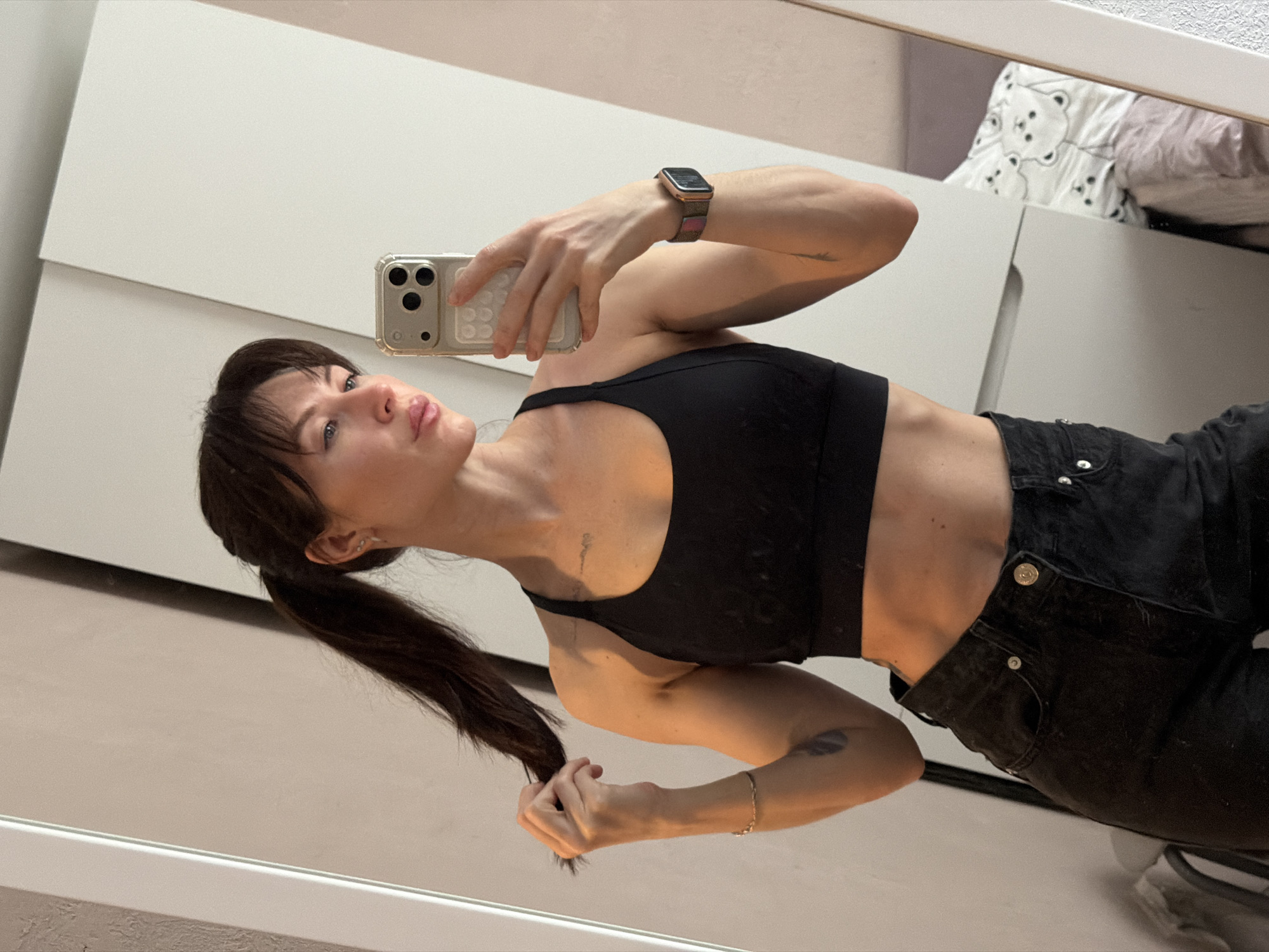 A young woman with dark hair in a ponytail is posing in a mirror selfie. She is wearing a black sports bra and black jeans. The background includes a white dresser. The location is indeterminate, and there is no specific evidence to indicate this is Perm, Russia.