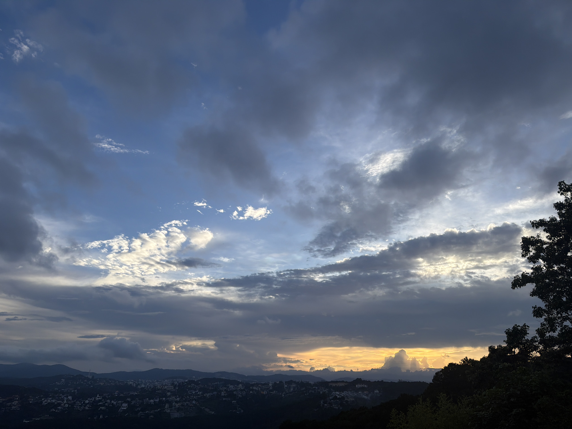 This is a wide-angle landscape photograph of a city nestled amongst rolling hills under a dramatic sky. The time of day appears to be late afternoon or early evening, as indicated by the warm, golden hues of the sunset breaking through the clouds. The weather is dynamic, with a mix of dark, heavy clouds and patches of bright, illuminated sky.
The foreground is dominated by the silhouettes of trees and lush foliage, creating a dark, textured border on the right side of the frame and extending across the bottom. Beyond the trees, a sprawling city is visible, its buildings rendered in dark tones against the slightly lighter landscape. The city appears to be built on hills, with structures cascading down the slopes. In the distance, layered mountain ranges create a sense of depth, fading into a hazy blue.
The sky is a prominent feature of the image, filled with a complex arrangement of clouds. Large, dark masses of clouds drift across the upper portion of the frame, contrasted by wispy, illuminated clouds that catch the light of the setting sun. These brighter clouds create a sense of texture and movement. The sun itself is not directly visible, but its light is suggested by the warm glow on the horizon and the illumination of the clouds. The overall mood of the image is one of natural grandeur and peaceful observation, with the human settlement appearing small and integrated within the vastness of the landscape and sky. There is no visible text in the image.