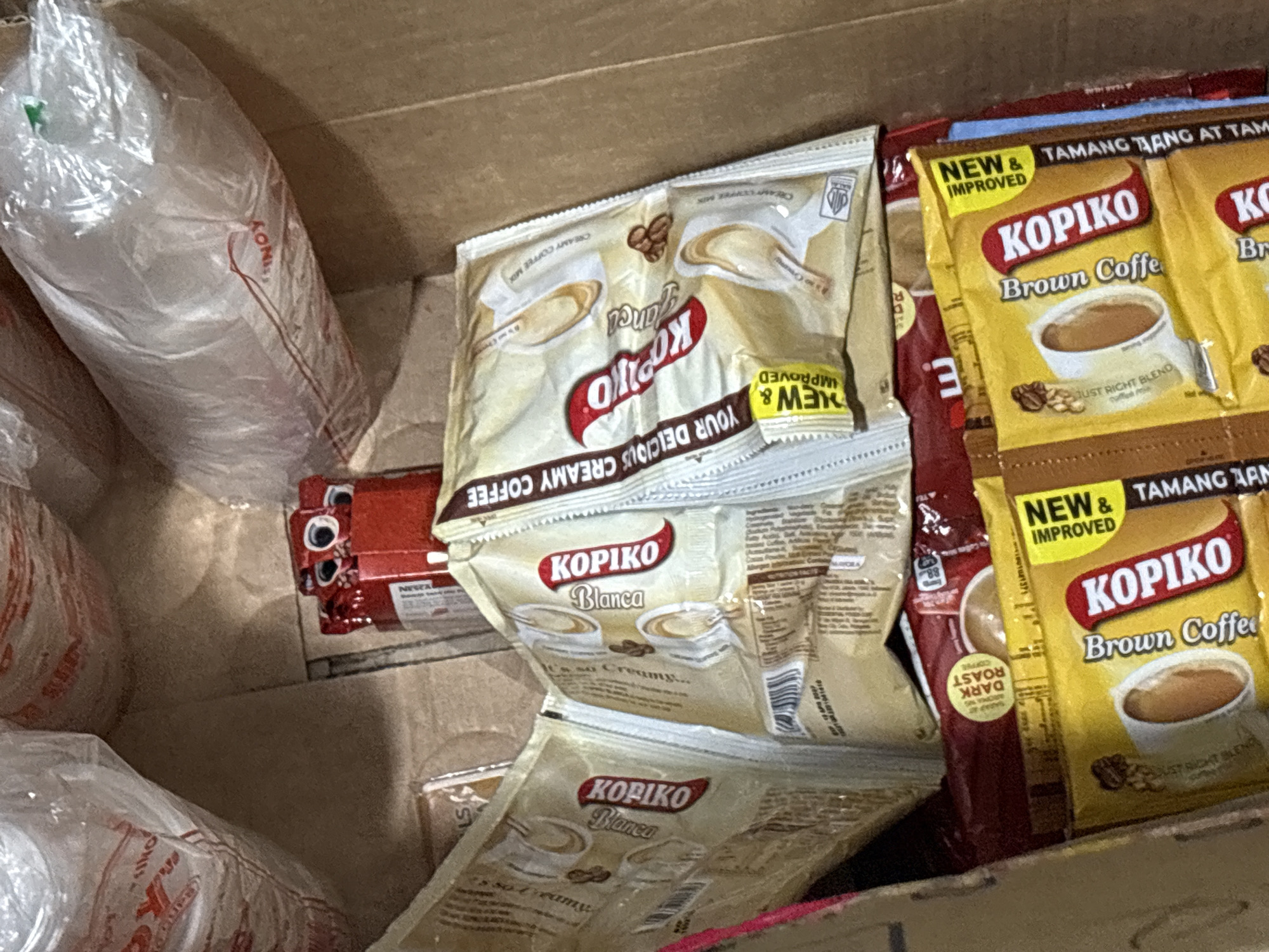 This image depicts the interior of a cardboard box filled with various packaged Kopiko coffee products. The items are stacked and arranged in a haphazard manner, suggesting they are either in storage or being prepared for distribution.
The majority of the visible items are individually wrapped sachets of Kopiko coffee. These include Kopiko Blanca, described as a "creamy coffee mix," and Kopiko Brown Coffee. Some sachets feature prominent branding, with the Kopiko logo in red and white letters, alongside images of coffee cups and beans. There are also some red-colored packets which are likely other variations of Kopiko coffee products.
In the background, there are large, opaque plastic bags, possibly containing more coffee products or related items. The overall environment appears to be an indoor setting, likely a storage area or a commercial space. The lighting suggests it is daytime. There are no people or animals visible in the image, and no clear indication of a specific location within Davao City, Philippines.