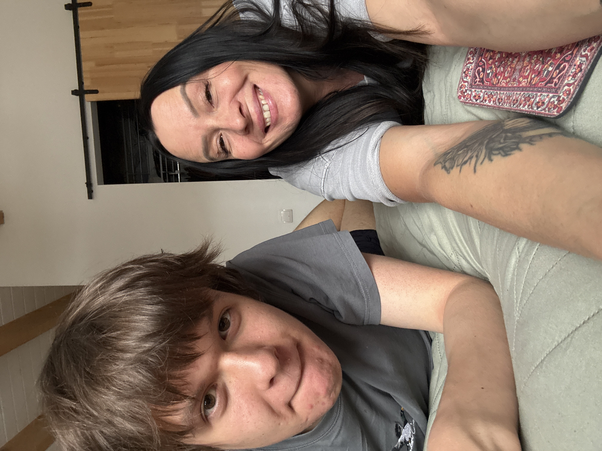 Two people are lying on a quilted surface, likely a bed or couch. One person with long, dark hair is smiling at the camera, wearing a grey shirt. The other person has brown hair and is also wearing a grey shirt. A tattoo is visible on the first person's arm. A patterned item, possibly a mousepad, is next to the woman. The location appears to be indoors in a residential setting. There is no information available in the image to confirm the location is Chaikovsky, Russia.