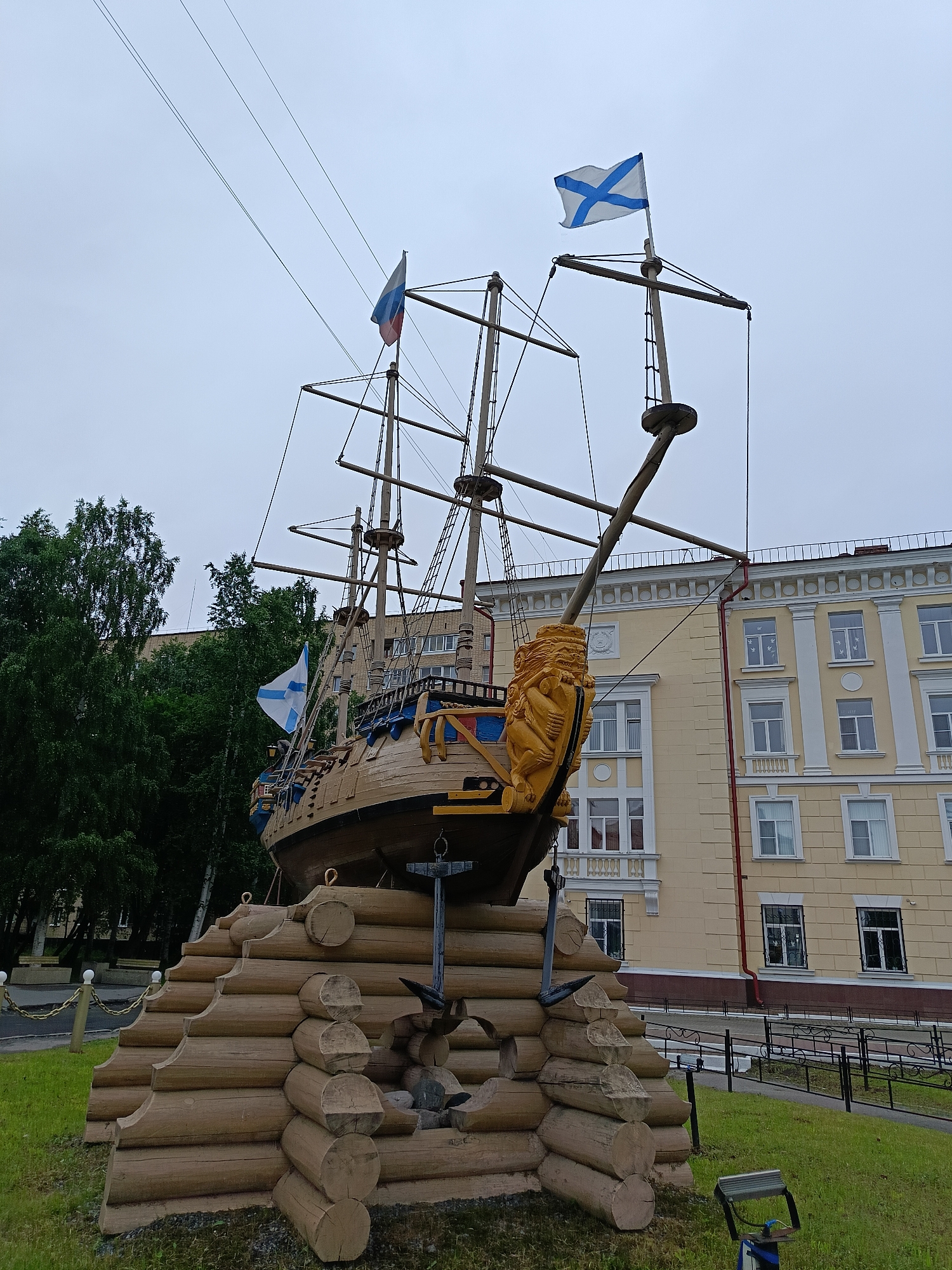 The image shows a sculpture of a wooden ship with three masts, adorned with flags and a golden lion's head at the bow. The ship is mounted on a tiered base constructed of logs, resembling a breakwater. Behind the sculpture stands a light-yellow building with white columns and numerous windows. Tall green trees are visible to the left of the building, and a grey, overcast sky fills the background. The scene is set in an outdoor area with a grassy lawn in the foreground. This monument is located in the city of Arkhangelsk, Russia.