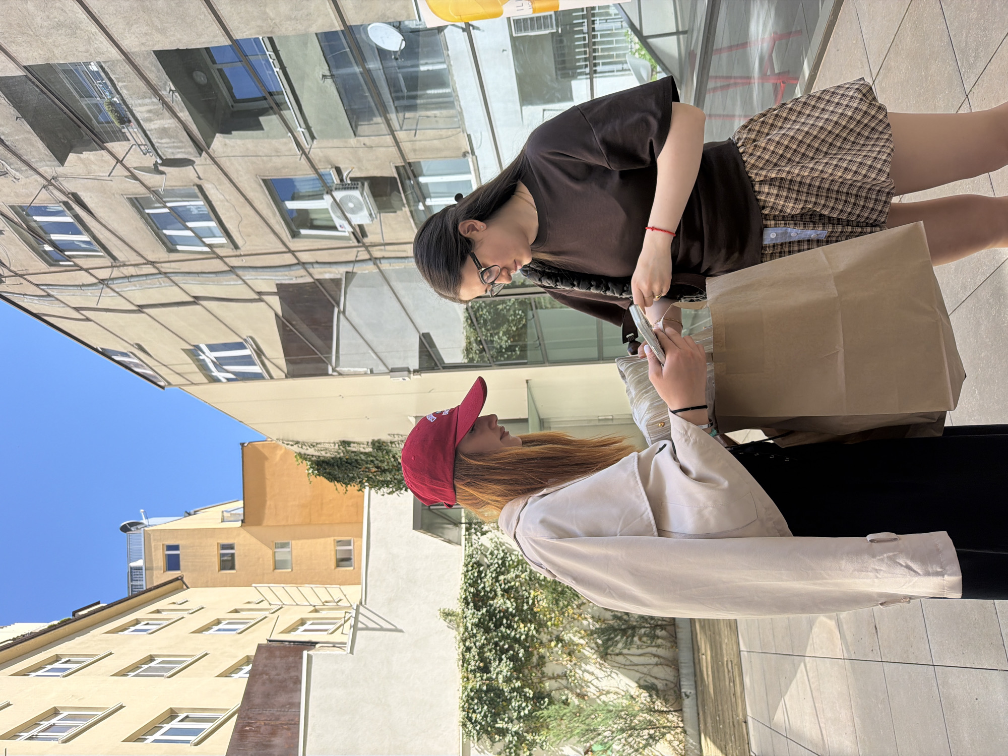 Two young women stand on a sidewalk in Sofia, Bulgaria. One woman wears a red baseball cap, a beige trench coat, and a black skirt. She is holding a smartphone. The other woman wears glasses, a brown t-shirt, and a plaid skirt and is holding a small item. Both are holding brown paper shopping bags. The buildings behind them reflect a mirrored image of the surrounding buildings. The sky is blue.