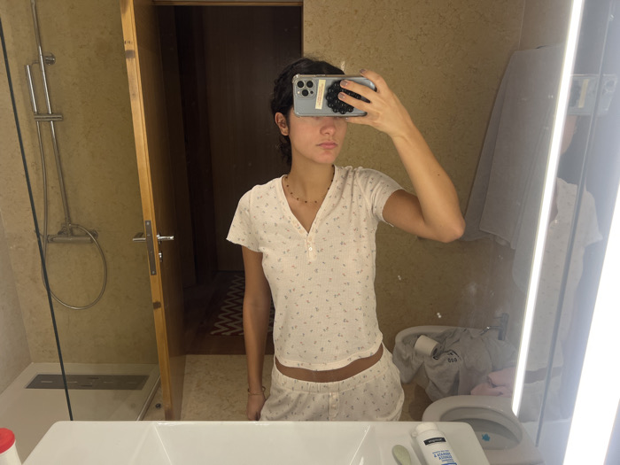 The scene is an interior view of a bathroom, located in Aldoar, Foz do Douro e Nevogilde, Portugal. A woman, captured from the chest to mid-thigh, stands centrally and takes a mirror selfie. She wears light-colored, short-sleeved pajamas featuring a small floral pattern, consisting of a V-neck henley top with buttons and matching shorts. A thin, dark beaded necklace is visible around her neck. Her right hand is raised, holding a smartphone (identified as an iPhone 14/15 Pro by its camera array) directly in front of her face, obscuring her facial features.
In the immediate foreground, a white sink basin is partially visible. On the counter to its left, a partial red cap is present. To its right, a white bottle labeled "Neutrogena 2 EM 1 Limpeza & Mascara" and a green applicator are positioned. The mirror itself incorporates two prominent vertical light strips along its right edge.
To the left of the woman's reflection, a modern shower area is visible, featuring a clear glass partition, beige tiled walls, a silver shower head, and a linear floor drain. Adjacent to the shower, a light-colored wooden door stands ajar, revealing a darker internal corridor with a patterned floor runner. To the right of the woman's reflection, a white toilet is visible, with a bidet faucet mounted on the adjacent wall. A pile of laundry, including a grey garment with a partially visible "ADIDAS" logo, rests on or near the toilet, alongside a roll of toilet paper. The bathroom walls are uniformly covered in beige-toned tiles or textured material.