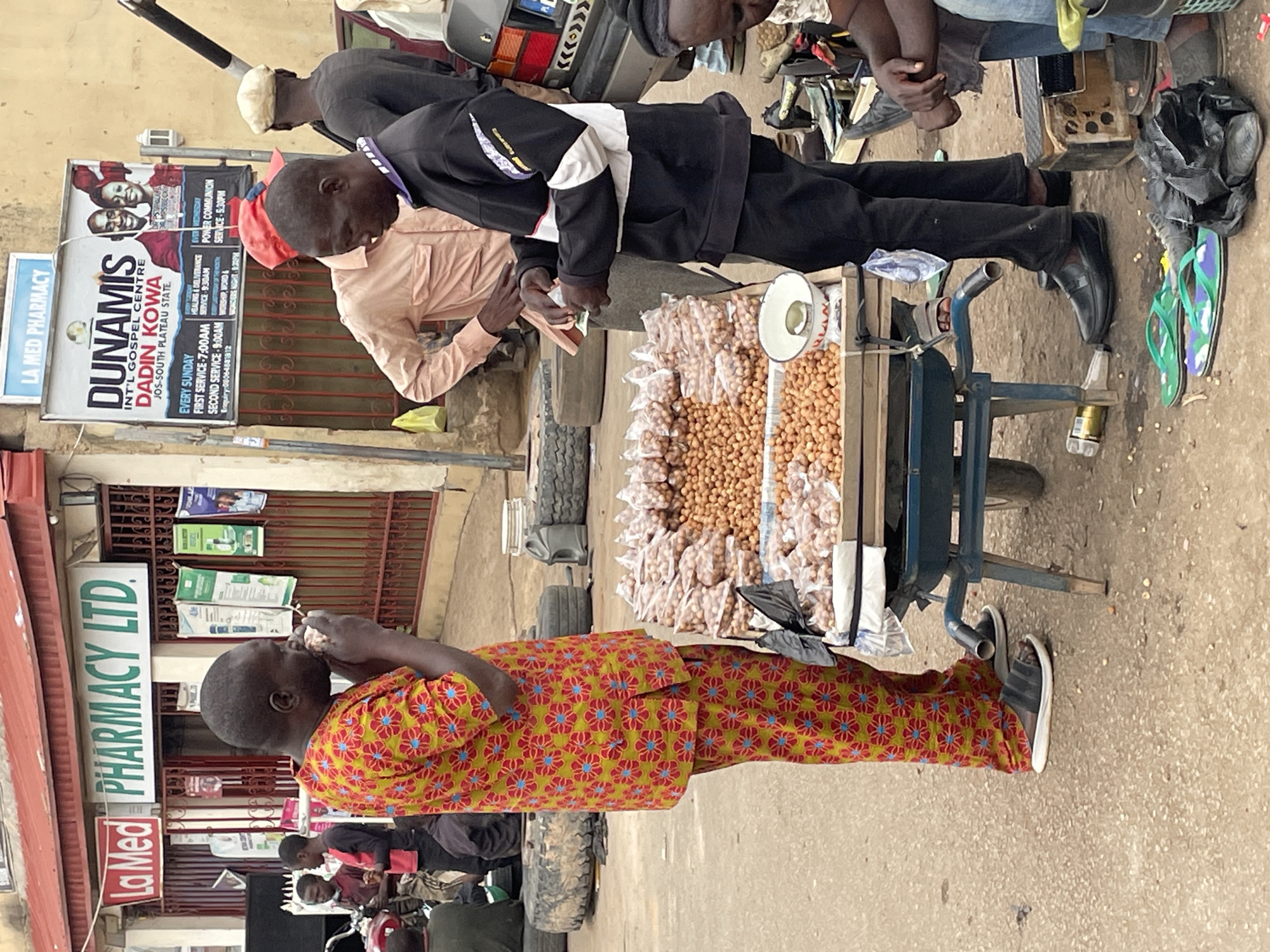 This image depicts a bustling outdoor street scene, likely a market or commercial area, in Jos, Plateau State, Nigeria, during daylight hours.
In the foreground, a young person or child stands wearing a vibrant, long-sleeved, patterned yellow, red, and blue traditional outfit, looking towards the right side of the frame while holding something to their mouth. They wear black sandals with white soles.
The central focus is a transaction occurring around a blue metal pushcart. The cart is filled with light brown, round items, resembling groundnuts or peanuts, some loose and some pre-packaged in clear plastic bags. A white measuring scoop or bowl is on the cart. A man in a light peach-colored long-sleeved shirt handles small items, possibly currency or goods. Beside him, another man, dressed in a black jacket with white and purple accents, black trousers, and black loafers, observes the interaction. In the background, other individuals, vehicle tires, and part of a gray car's rear bumper are visible. Green and purple flip-flops lie on the ground next to the pushcart.
The immediate surroundings feature several prominent signs. A large sign for "LA Med PHARMACY LTD." with green lettering is visible, along with a smaller "LA MED PHARMACY" sign in red. A prominent banner advertises "DUNAMIS INT'L GOSPEL CENTRE DADIN KOWA JOS-SOUTH PLATEAU STATE." Details on the banner include service times: "EVERY SUNDAY FIRST SERVICE - 7:00AM," "SECOND SERVICE - 9:00AM," and "HEALING & DELIVERANCE SERVICE: 9:30AM." It also lists "EVERY WEDNESDAY POWER COMMUNION SERVICE - 5:30PM," and a partial enquiry number: "0006-4881812," alongside "WORSHIP, WORD & WONDERS NIGHT-120."
The scene captures a moment of daily commerce and public life under bright, clear weather conditions, characteristic of an urban environment in Nigeria.