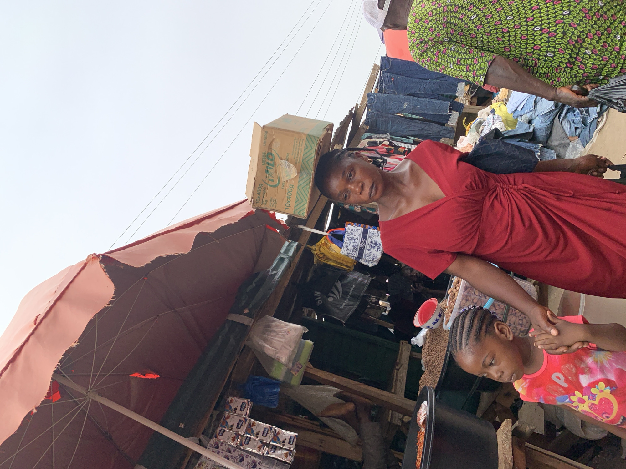 This image captures a vibrant, sunlit market scene in Akwanga, Nigeria, showcasing daily life under a bright, clear sky. The main subjects are a woman in a red dress and a young girl, who are standing near a market stall. The woman, with braided hair, looks directly at the camera with a neutral expression, gently holding the hand of the young girl. The girl, wearing a pink and yellow patterned t-shirt and with braided hair, looks downwards, seemingly focused on something below. An older person, partially visible in a green patterned top, stands to the right, observing the scene.
The market stall is sheltered by a large, somewhat worn red umbrella or awning. The structure of the stall appears makeshift, constructed with wooden planks, and is filled with various goods. Numerous pairs of jeans and other clothing items are displayed, hanging behind the people. A prominent "Nestle MILO" cardboard box is visible, displaying details such as "10x400g", "Net weight: 4kg", and "NAFDAC Reg.NO 01-0070". Other market items include packaged goods, plastic bags (one with the text "Bag for recycling" and another possibly "SIN"), and containers holding foodstuffs like peanuts and a dark stew in a black pot. A white plastic bowl with "AK WA" (likely part of "Akwanga") written on it is also seen. The setting suggests a busy, open-air market environment.