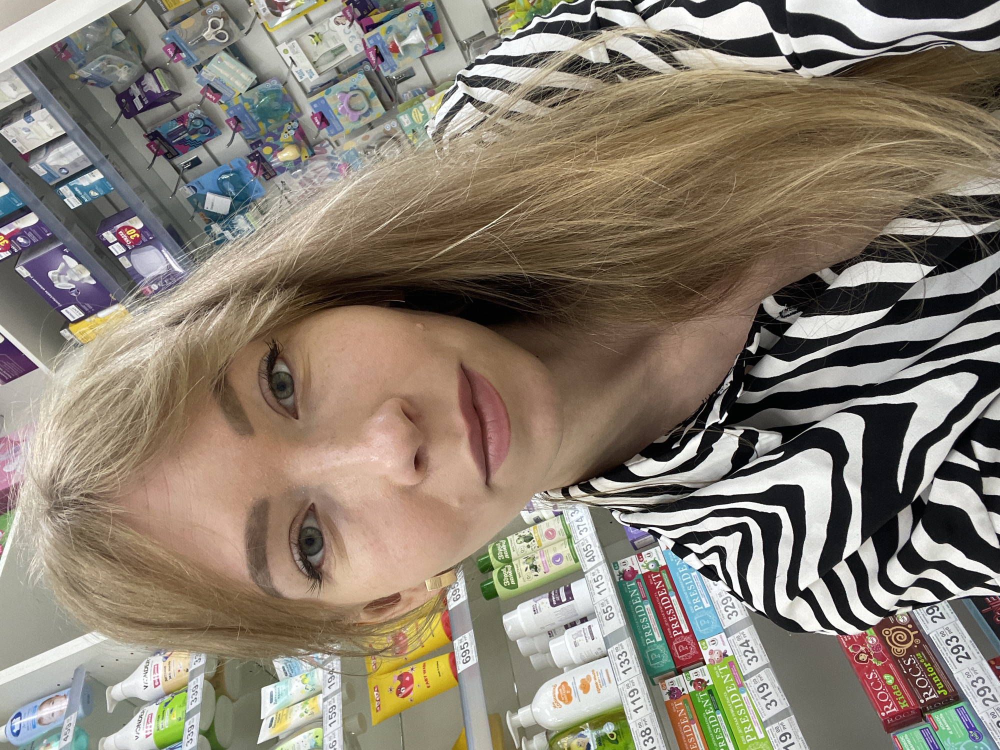 The image shows a young woman with blonde hair and green eyes taking a selfie in what appears to be a pharmacy or a store selling baby products. She is wearing a black and white zebra-print shirt. In the background, shelves are stocked with various products, including bottles of baby wash, toothpaste, and other personal care items. Price tags are visible on some of the shelves. The scene is well-lit, suggesting an indoor setting. The woman's face is the primary focus, with her hair falling over her shoulder. The location is identified as the city of Perm, Russia.