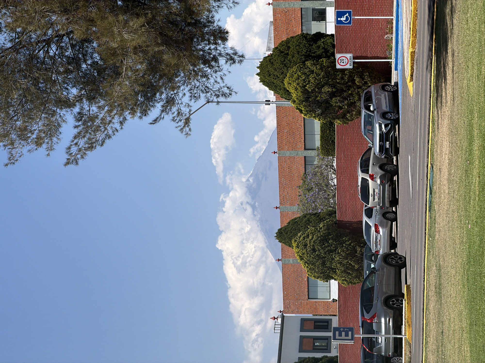 The image depicts an outdoor scene in San Andrés Cholula, Mexico, during daylight hours. A clear blue sky, interspersed with white cumulus clouds, dominates the background. The peak of a large, distant mountain, possibly Popocatépetl or Iztaccihuatl, is visible through the clouds.
In the midground, a red brick building features green decorative elements and windows. Lush green shrubs and trees, some with purple blossoms, are planted around the building. A tall streetlamp extends upwards near the center.
The right side of the frame is occupied by a paved parking area with yellow and blue painted lines. Several cars, including a grey SUV and a silver sedan, are parked in a row. Two prominent signs are visible on white poles: a blue sign with a white wheelchair symbol designates accessible parking, and a circular sign indicates a "20 MAXIMA" speed limit. Further to the bottom right, a white square sign with a blue 'E' and an arrow likely denotes parking. Green grass borders the parking lot on the far right. The upper left corner features the dark, needle-like foliage of a large tree. No people are present in the image.