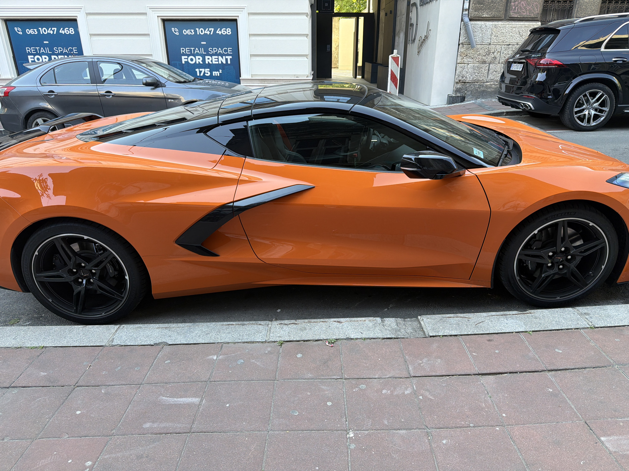 The image shows a bright orange Corvette coupe parked on the sidewalk next to a brick-tiled street in Belgrade, Serbia. The car has black accents, including the roof, side air intakes, and wheels. Behind the Corvette, there is a dark gray sedan and a black SUV parked on the street. In the background, there are two store windows with blue signs that read "063 1047 468 RETAIL SPACE FOR RENT 175 m²" and "063 1047 468 RETAIL SPACE FOR." The building facade is white with a dark entryway.
