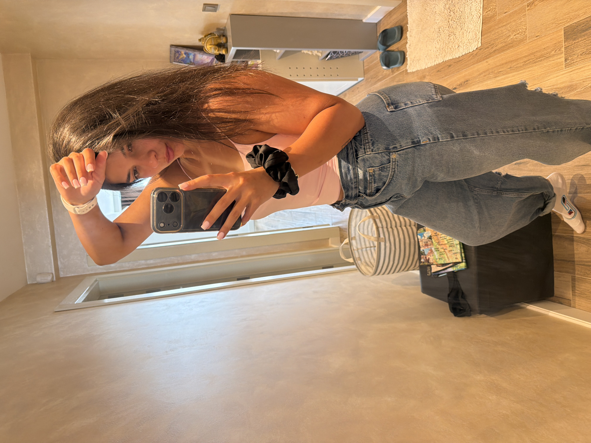 A young woman with long, dark hair is taking a mirror selfie in a hallway. She is wearing a light pink tank top and loose-fitting light wash denim jeans with ripped details. She has a black scrunchie on her left wrist and a white wristband on her right wrist. Her hand is resting on her forehead. In the background, there is a large mirror on the left, a doorway, a shelving unit with decorative items, a hamper, a dark bench with magazines and a black mask, and a pair of blue flip-flops on the floor. The flooring is wood-patterned. The overall scene suggests a domestic or hotel room setting, not a location in Gabrovo, Bulgaria.