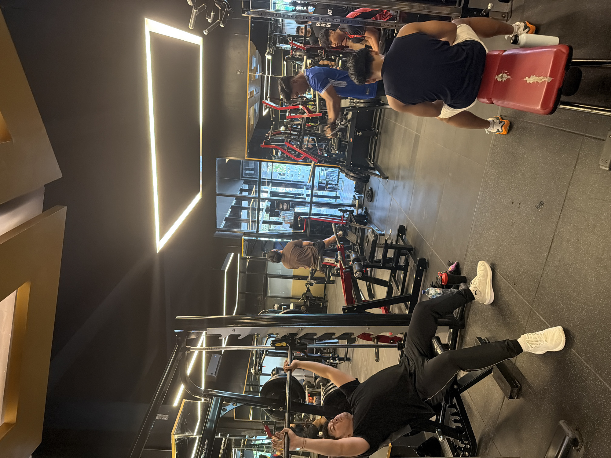 The image depicts the interior of a gym located in Denpasar, Indonesia. Several individuals are engaged in various exercises. In the foreground, a person lies on a weight bench, lifting a barbell within a Smith machine. In the midground, a person is seated on a weight machine. Further back, near a mirrored wall, two individuals are using other exercise machines. The gym is equipped with standard weightlifting equipment and has a modern aesthetic with black walls and linear LED lighting fixtures. Natural light enters through large windows.