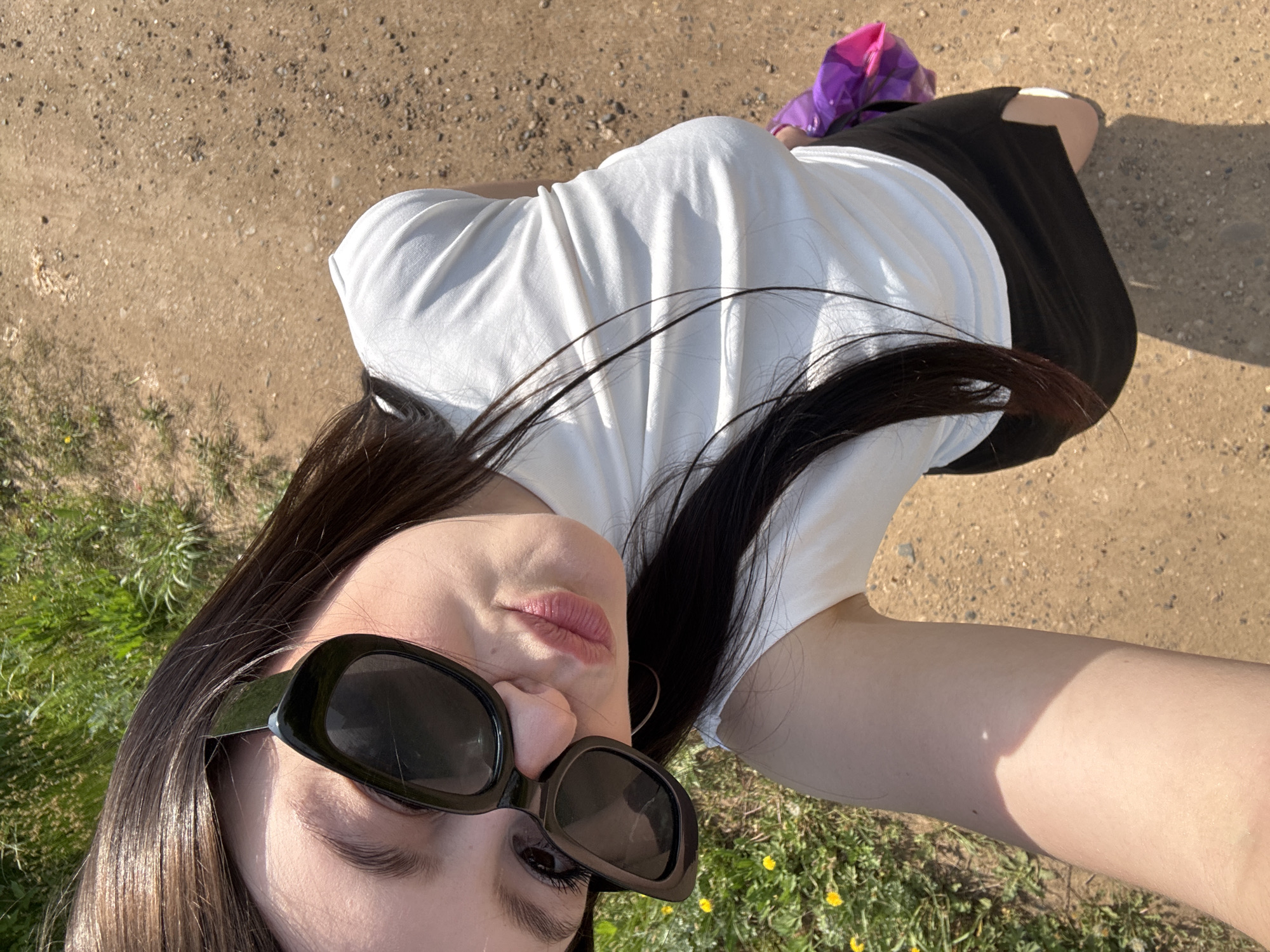 A young woman wearing sunglasses, a white shirt, and a black skirt is lying on the ground in what appears to be a park or natural area. The ground consists of dirt and patches of green grass with small yellow flowers. The area appears to be Фоки, Russia. A purple object is visible next to her skirt.