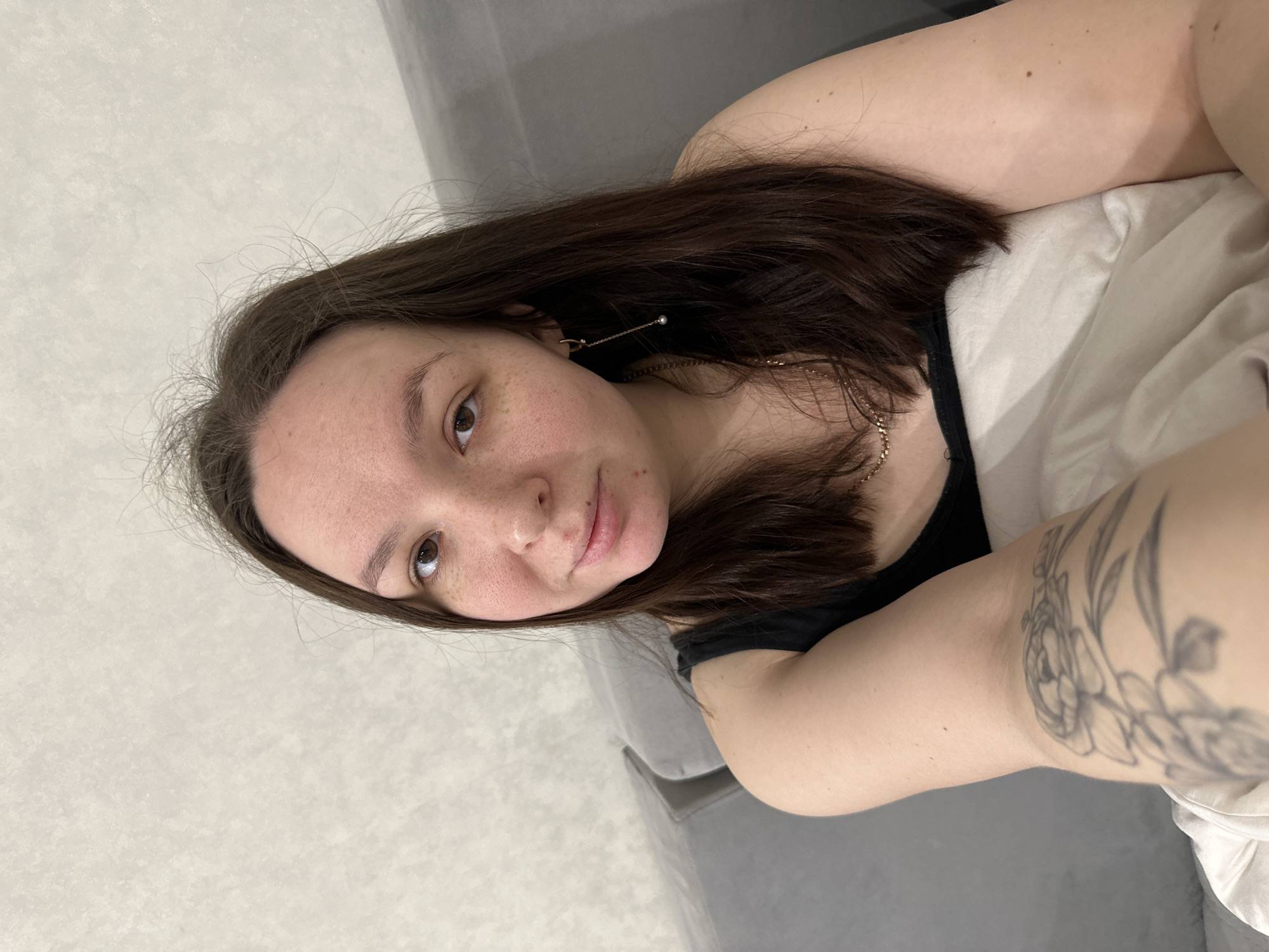 The image depicts a young woman in an indoor, residential setting, presumed to be within the city of Chaikovsky, Russia. The woman, who appears to be in her late teens or early twenties, has medium-long, dark brown hair, parted to the viewer's left. Her skin is light-toned with visible freckles primarily on her nose and cheeks. She has brown eyes and a slight smile, looking directly at the camera. She is wearing a dark-colored tank top or camisole. A gold-colored chain necklace is visible around her neck, and a gold-colored dangling earring with a small pearl hangs from her right ear (viewer's left). A floral tattoo, depicting what appears to be roses, is present on her left upper arm. She is seated or reclining on a grey upholstered sofa or similar furniture, part of which is visible behind her to the viewer's right. The background consists of a light-colored, textured wall. A white or light-colored blanket or pillow covers her lower body and is held by her left arm. The scene is illuminated by ambient indoor light.