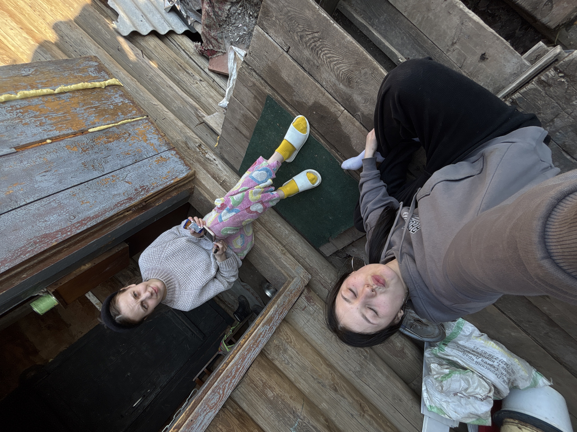 Two women are positioned on a weathered wooden surface that appears to be an outdoor deck or porch. One woman, situated in the lower left portion of the image, is wearing a light gray sweater, patterned pajamas with pink, yellow, and blue designs, and yellow socks with white slippers. She is holding a small rectangular object in her hands, possibly a phone or cigarette. The other woman, in the upper right of the image, is wearing black pants, a gray hoodie, and a white sock on one foot. Her eyes are closed, and she appears to be taking a selfie. A bag with Cyrillic text is near her, with the visible text suggesting the location is within the Perm Krai region of Russia, specifically Фоки (Foki). The wooden surface surrounding the women is made of planks in various states of wear, with some areas showing decay or damage. In the upper left corner, corrugated metal sheets are visible. The overall environment suggests a rural or semi-rural setting in Фоки, Russia.