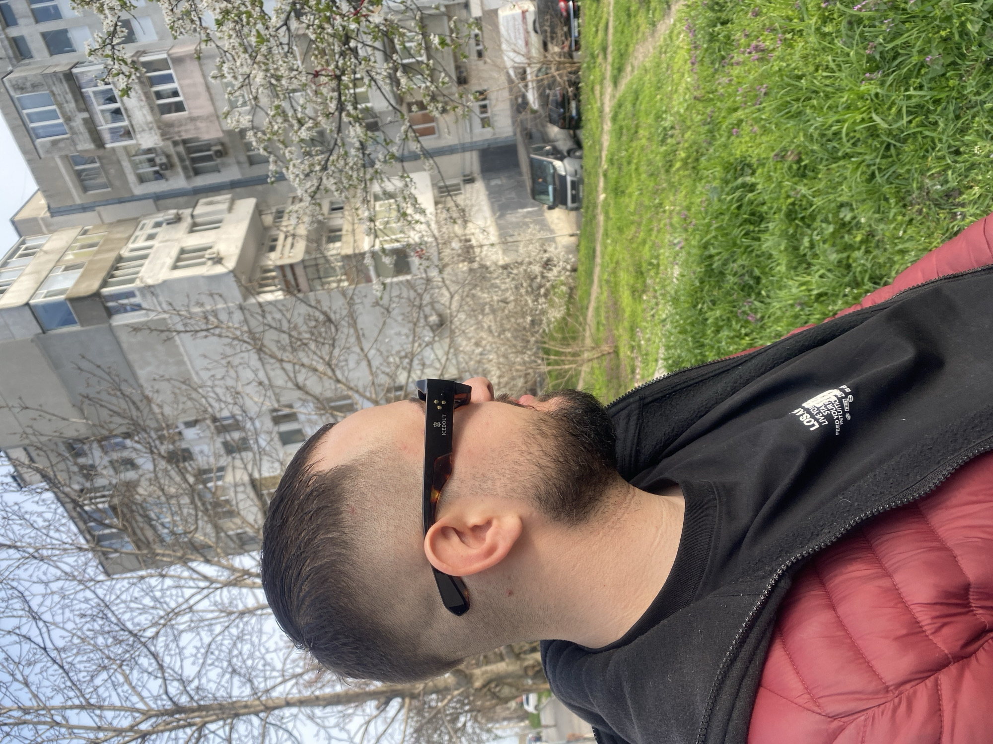 A medium shot depicts a man wearing sunglasses. He is seen from the shoulders up, turned to the left. He has short, dark hair, a beard, and is wearing a black t-shirt with white writing and a red quilted jacket. He is wearing sunglasses with dark frames.
The background consists of a concrete building, a blossoming tree, and a grassy area with some purple flowers. Based on the architecture and surroundings, this image was likely taken in a residential area of Varna, Bulgaria.