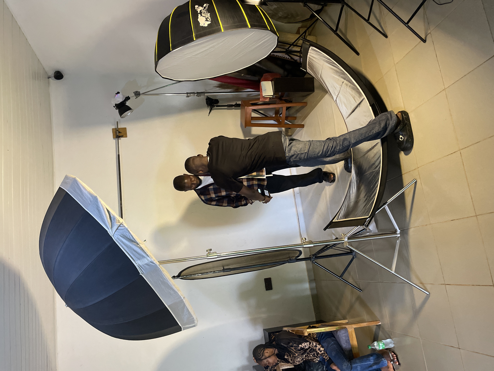 In Durumi, Nigeria, a photography studio is set up for a portrait session. Two men are the subjects, with one man standing and the other in front of him appearing to adjust his shirt. A woman is seated on a chair in the background. The studio is equipped with professional lighting equipment, including a large umbrella light, a softbox, and a reflector. A stool and a few other items are also present in the room. The floor is tiled, and the walls are white.