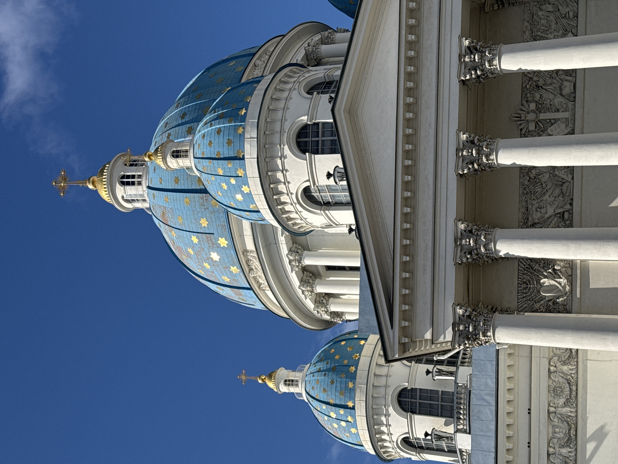 This upward-angled photograph showcases the grand architectural details of a cathedral against a vibrant, clear blue sky under bright daylight. The primary subjects are two magnificent light-blue domes, adorned with numerous golden stars of varying sizes. Each dome is crowned with a white, multi-windowed drum, a golden onion-shaped cupola, and a golden Orthodox cross at its apex.
Below the domes, a section of the cathedral's white or light-colored stone facade is visible. This facade features classical columns with intricately carved Corinthian capitals. Between and above these columns, detailed bas-reliefs with religious and symbolic motifs are carved into the stonework, depicting various scenes and elements like crosses, angels, and foliage. Arched windows are also discernible on the lower sections of the building.
The bright sunlight casts distinct shadows, highlighting the three-dimensional depth and ornate nature of the architectural elements. The weather appears clear and sunny, with only a few faint clouds visible in the expanse of the deep blue sky. No people are visible in the frame, and there is no discernible text. The striking color contrast between the light-blue domes, golden accents, and the white stone against the expansive sky emphasizes the grandeur and beauty of the structure. This architecture is characteristic of the Trinity Cathedral (Troitsky Sobor) in Saint Petersburg, Russia.