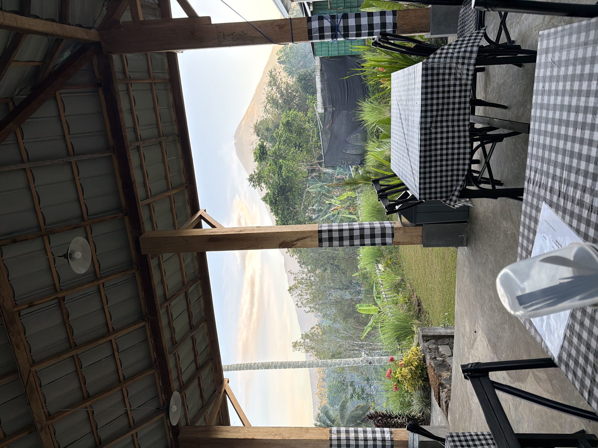 An open-air covered patio overlooks a lush green landscape with a prominent mountain in the distance, illuminated by early morning or late afternoon light. Two wooden pillars with black and white checkered fabric draped around them support the corrugated metal roof. In the foreground, two tables are set with black and white checkered tablecloths and black chairs. One table has a clear plastic cup containing utensils placed on a menu. The background features dense foliage, including banana trees and a tall, slender palm tree, with a stone wall and neatly mown grass. A black tarp covers a structure behind some of the vegetation. The overall scene suggests a restaurant or cafe situated in a rural area, likely in Sidomulyo, Indonesia, known for its scenic mountainous terrain.