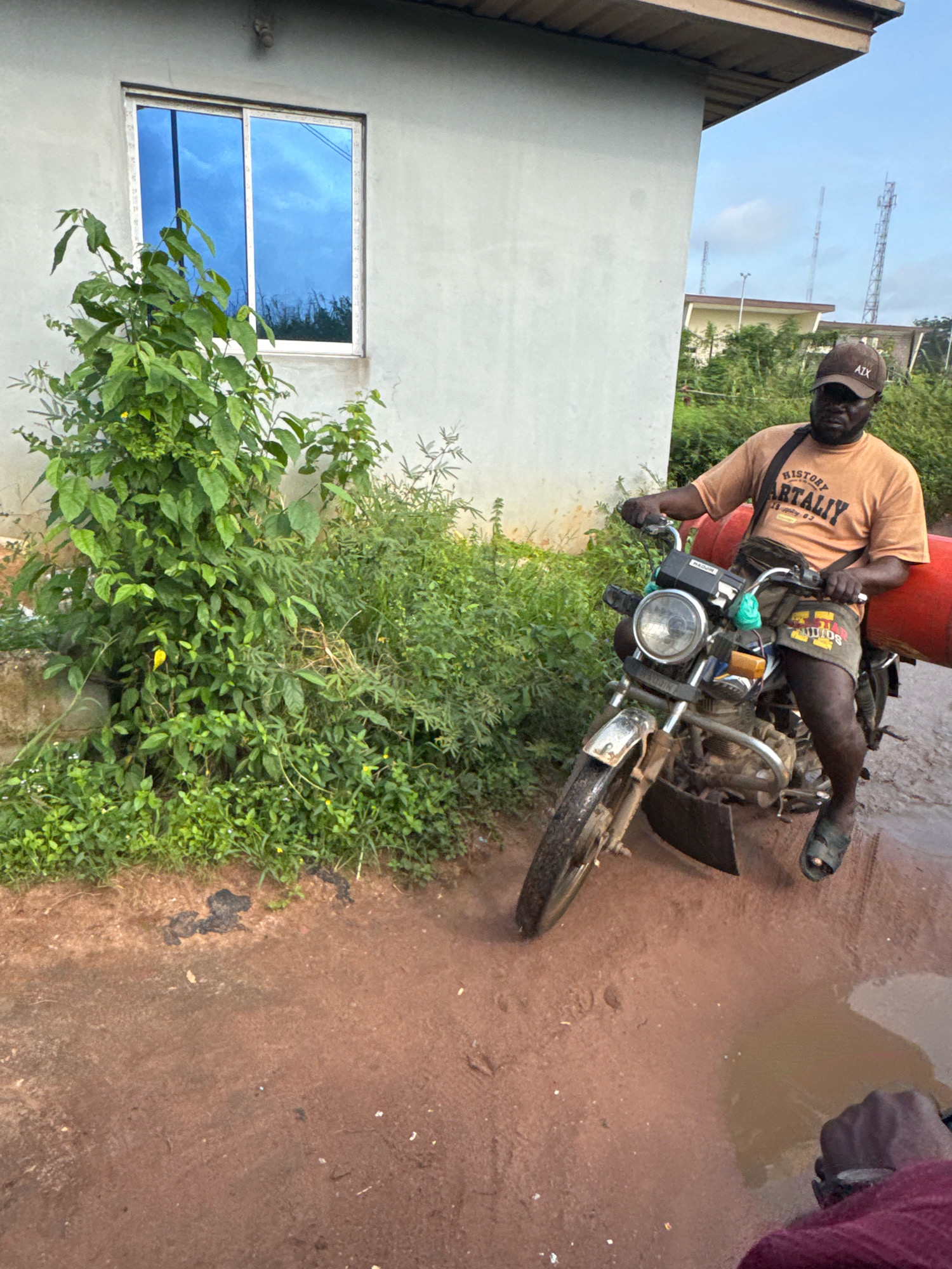 A dark-skinned man wearing a tan t-shirt with "HISTORY ARITALIY" printed on it and a dark brown baseball cap is sitting on a dirt-covered motorcycle in a muddy, reddish-brown area. He is looking forward and has a neutral expression. The motorcycle has a red cylindrical object strapped to the back, extending over the man's right shoulder. He is wearing patterned shorts and dark sandals. The motorcycle's front wheel is slightly turned to the left. In the background, there is a light-colored building with a blue-tinted window. To the left of the motorcycle, lush green bushes and plants grow near the building. The ground is uneven and appears to have puddles of water. The sky is visible above, with some clouds and what appear to be communication towers in the distance, suggesting an outdoor, possibly rural or suburban, setting in Ihiagwa, Nigeria.