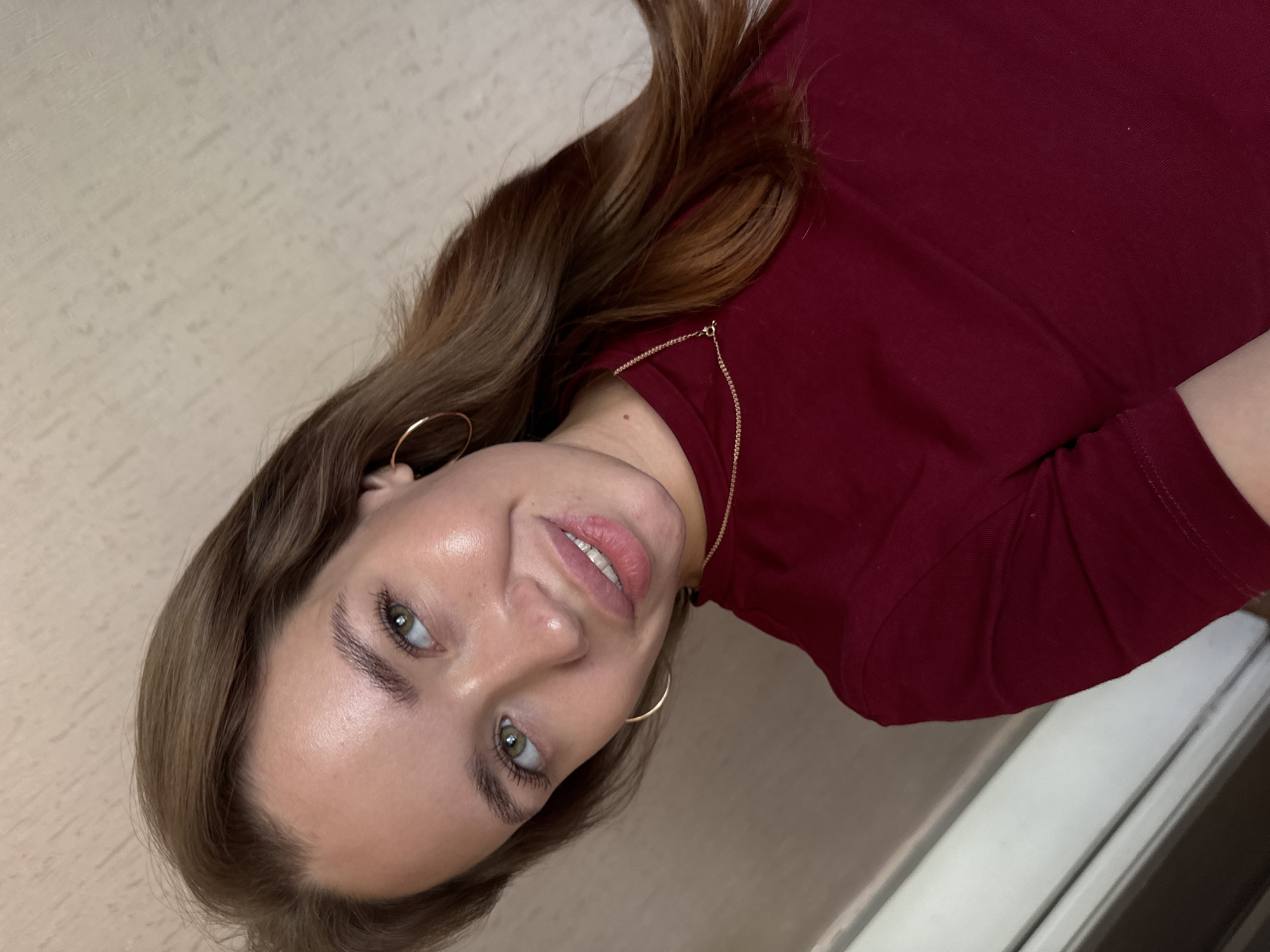 A young woman with long, brown hair is looking towards the camera, her head tilted slightly to the left. She is wearing a maroon-colored shirt and a delicate gold chain necklace. Her makeup includes mascara and lip gloss, and she is wearing hoop earrings. Her skin appears to have a dewy finish. The background is a neutral-toned wall with a textured pattern. The lighting suggests an indoor setting. There is no discernible information in the image to indicate a specific location in the city of Chaikovsky, Russia.