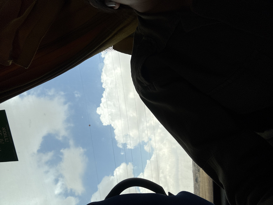 The image shows the interior of a bus, taken from the perspective of someone seated near the window. The window is open, revealing a view of the sky with clouds and a small, red object. The sky is visible through a window in the bus, with a cloudy sky and power lines overhead. The viewer's hand is visible in the foreground, and their torso and the seat beside them are obscured by their silhouette. The bus is travelling through a rural area. A sticker reading "אסור לה | NO | Smoking" is stuck on the inside of the window. The GPS coordinates place this location in Rahat, Israel. The image was taken during the day. The sky is bright, and the clouds are white. The landscape appears to be barren.
