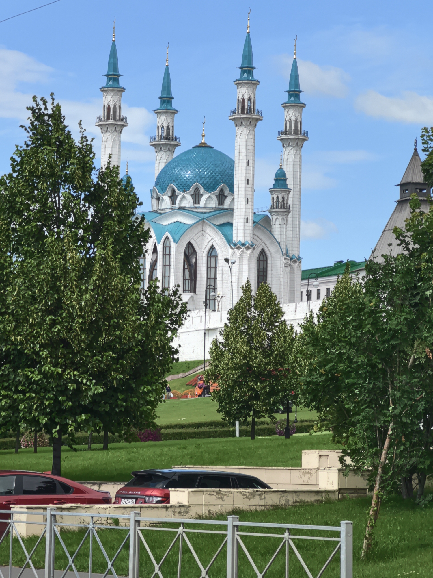 The image depicts a view of the Kul Sharif Mosque in Kazan, Russia. The mosque's prominent features include its white facade, turquoise domes, and tall, slender minarets topped with crescents. Lush green trees in the foreground partially obscure the mosque, with some branches and leaves creating a natural frame for the building.
In front of the mosque, a well-maintained lawn slopes gently upwards. A low, light-colored wall separates the foreground from the middle ground, where a red sedan and a red SUV are parked. The SUV has a license plate that reads "T243EP716". Beyond the cars and wall, the green lawn is visible, with some flowerbeds adding pops of color in the distance.
The sky above is a clear, bright blue with scattered white clouds, suggesting fair weather. The lighting indicates it is daytime, likely late morning or early afternoon, with the sun casting soft shadows. The scene is tranquil and serene, highlighting the architectural beauty of the mosque and its surrounding park-like environment. The overall impression is one of peaceful contemplation and architectural grandeur.