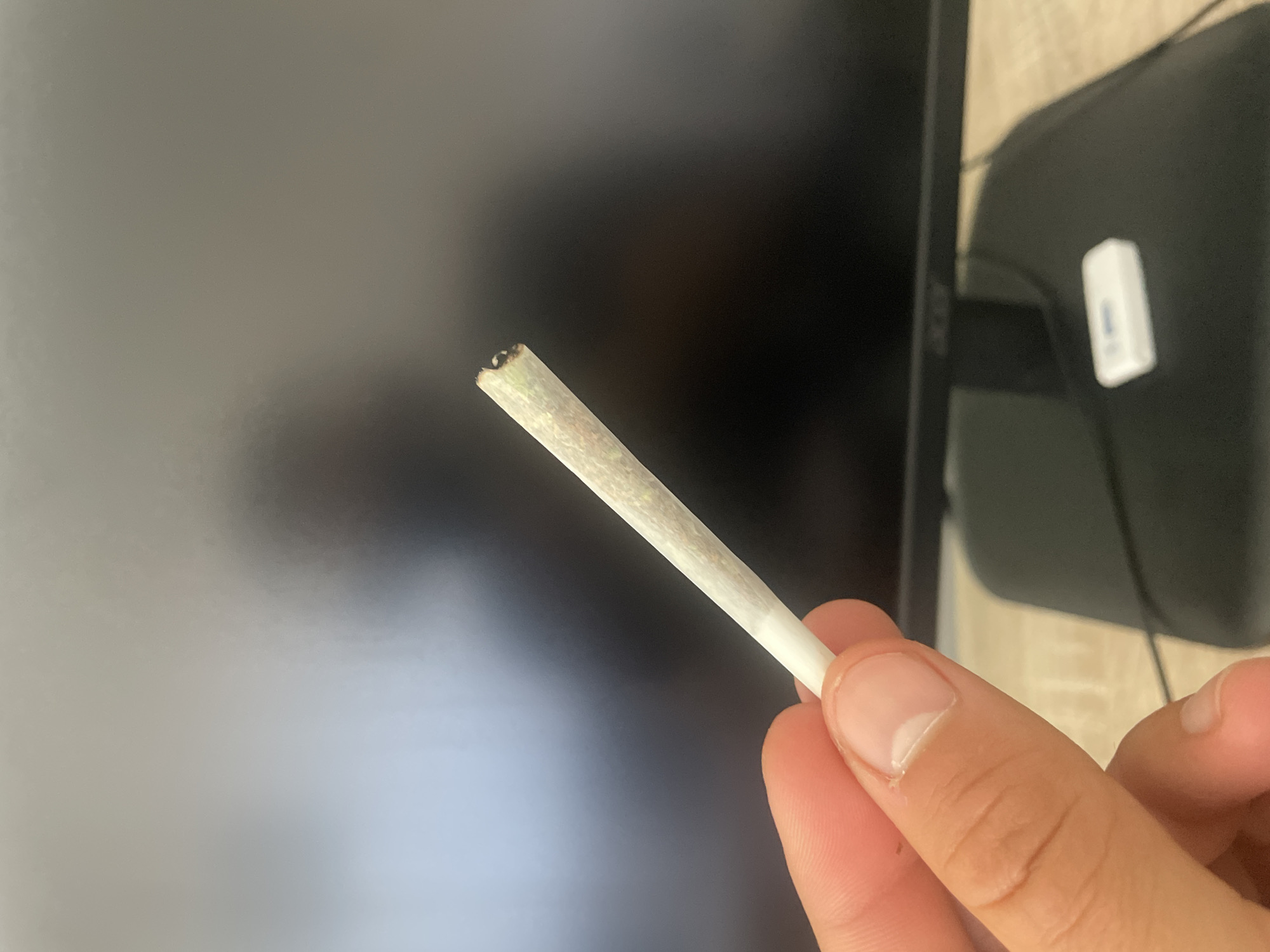 In an indoor environment, a close-up shot focuses on a person's hand holding a hand-rolled cigarette. The cigarette is positioned diagonally, extending from the lower left to the upper right of the frame. The hand, with visible fingernails and skin texture, grips the cigarette near its base. The cigarette itself appears to be made of light-colored paper, with a visible filter tip. The background is dominated by a dark, out-of-focus monitor screen, with a lighter area on the left suggesting a light source. Below the monitor, a blurred desk surface and the base of the monitor stand are visible. A small white rectangular object, possibly a USB drive, is present on the desk. The lighting is somewhat dim, with the primary light source illuminating the hand and cigarette.