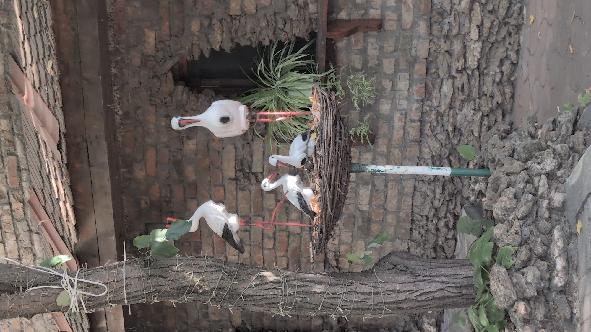 The image depicts a decorative stork nest with four stork figures situated outside a brick building with a tiled roof. The nest is supported by a pole and contains three adult stork figures, two of which are in the nest itself, and another standing tall behind them. A fourth, larger stork figure stands to the right of the nest. The background features a brick wall with a partially visible window and some greenery. A tree trunk adorned with string lights is visible on the left side of the image. The ground is covered with stones and some fallen leaves. The scene appears to be outdoors, possibly in a park or a decorative area within the city of Izmail, Ukraine. There are no people or animals present.