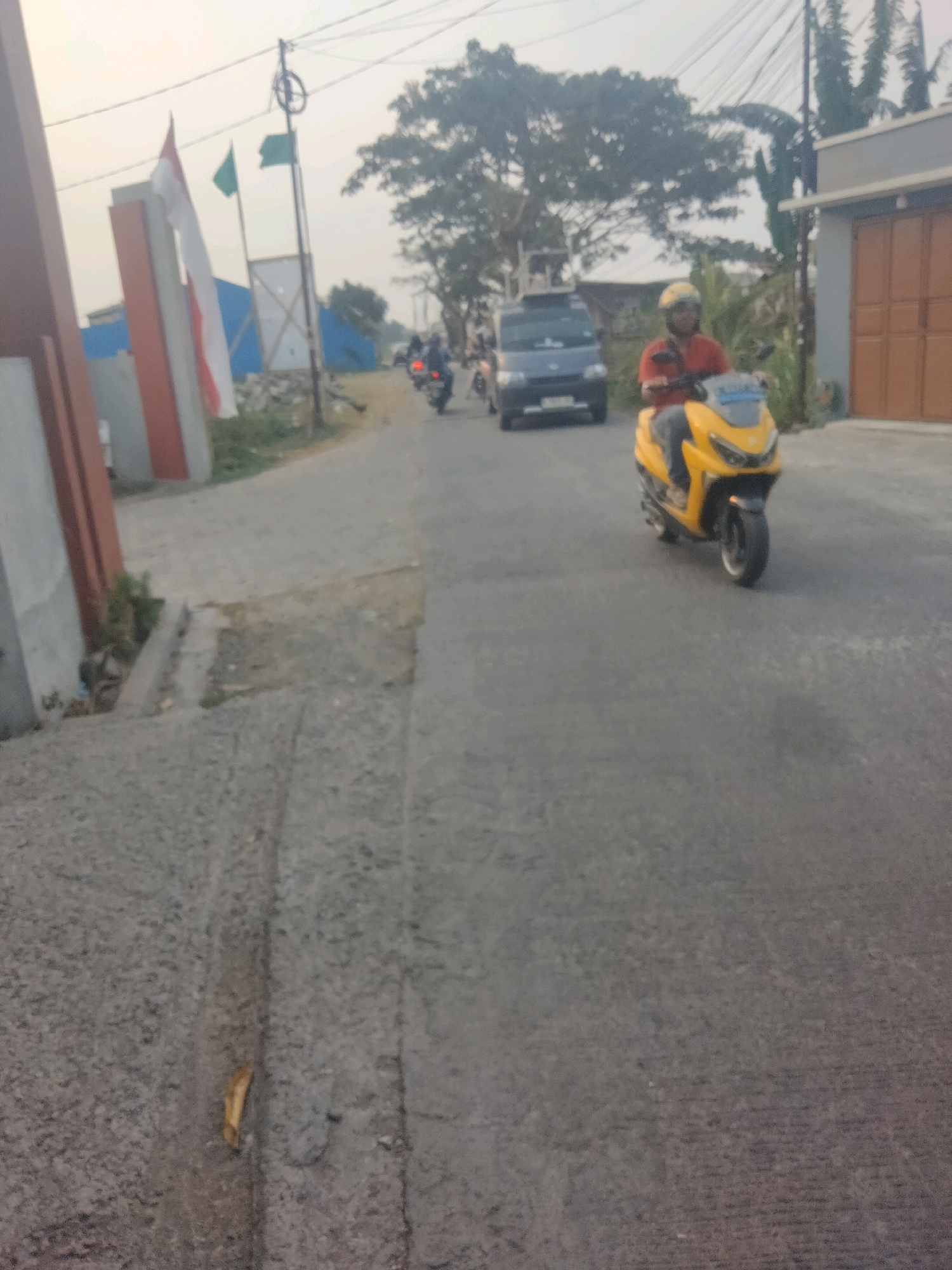 The image depicts a street scene in Suka Sari, Indonesia, during the day. A person wearing a red shirt and a yellow helmet is riding a yellow scooter. The scooter is a modern, sporty model. In the background, a dark-colored van is visible, suggesting traffic. There are also Indonesian flags displayed on poles, indicating a possible national event or location. The road surface is asphalt, and the surrounding environment includes buildings and trees. The lighting suggests it is either early morning or late afternoon. The overall atmosphere is that of a typical day in a semi-urban or suburban area.