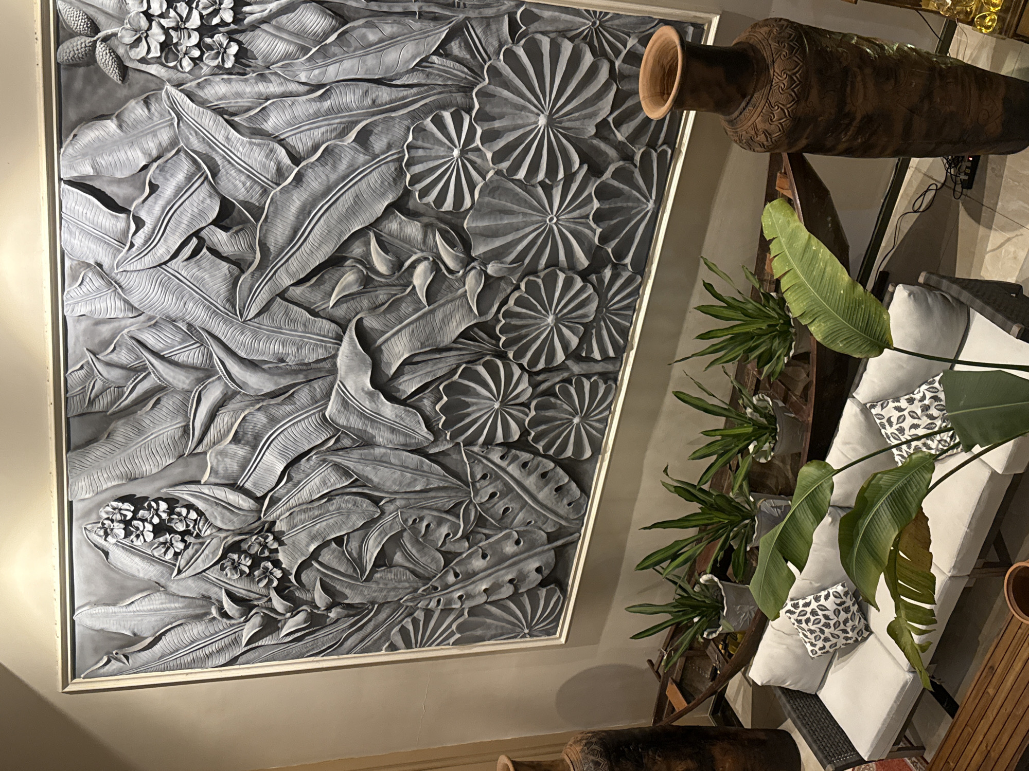 The image showcases a striking wall relief featuring a dense arrangement of tropical leaves and flowers, rendered in a muted gray tone. The relief is framed and mounted on a light-colored wall. To the right of the relief, a large, ornate, dark brown ceramic vase is positioned on a stand, its opening facing upwards. Below the vase and extending towards the foreground, a sectional sofa with white cushions and black and white patterned accent pillows is visible. Several potted green plants with large leaves are placed around the sofa area. A wooden slatted surface, possibly part of a table or bench, is also present on the right side of the image. The scene appears to be an indoor setting, possibly a living room or entryway, with artificial lighting contributing to the overall ambiance. No people are visible in the image. The style suggests a tropical or bohemian decor.