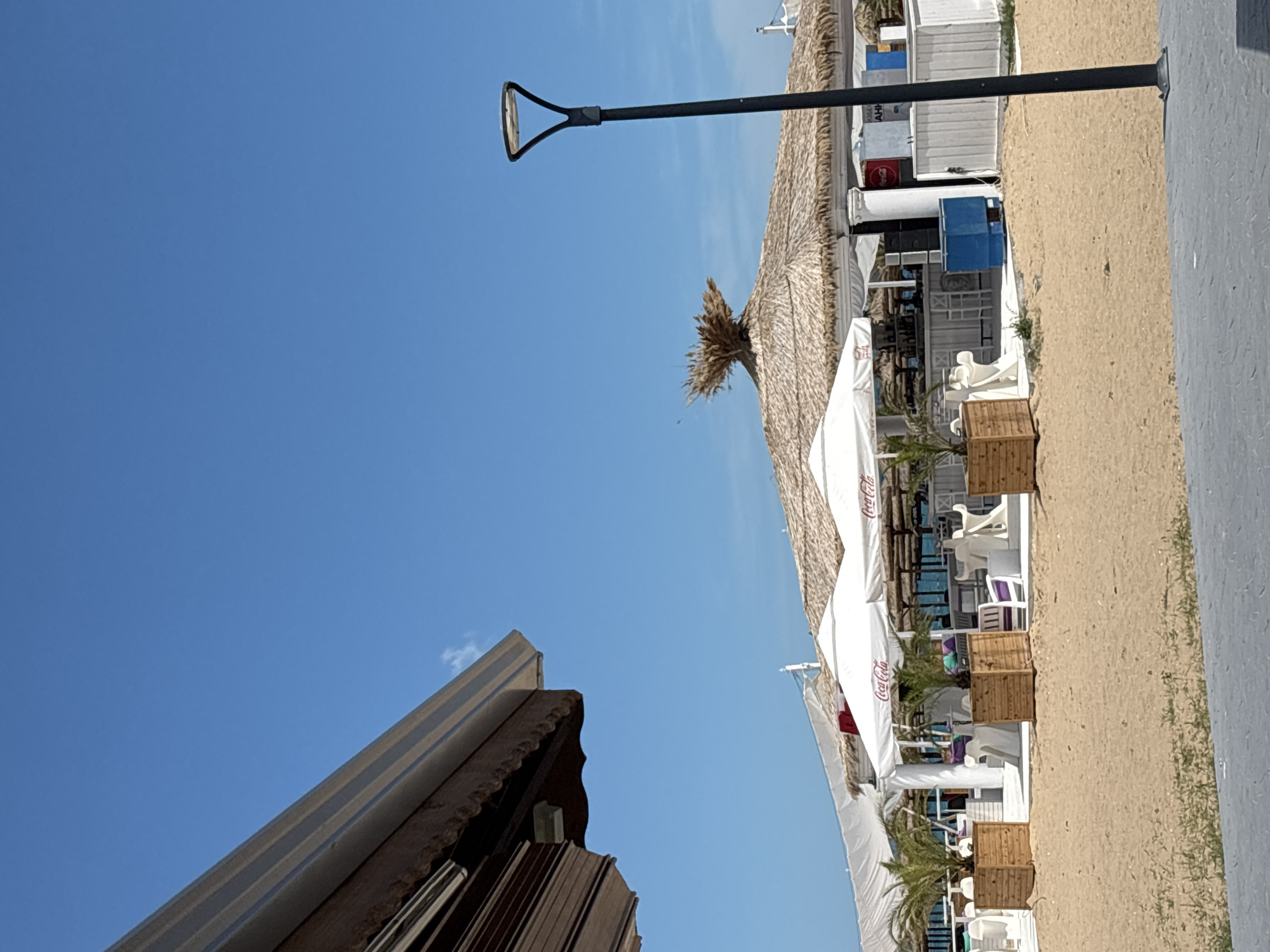 The image shows a beach bar area in Varna, Bulgaria, on a sunny day with a clear blue sky. The foreground features a sandy beach and a gray paved area. In the midground, there are several white umbrellas and a thatched roof structure that appears to be a bar or restaurant. Wooden planters with small palm trees are placed in front of the umbrellas. White chairs and tables are visible, suggesting an outdoor seating area. The background shows the sea, though it is mostly obscured by the structures. A black lamppost stands to the right of the image. The overall scene depicts a relaxed, outdoor establishment likely catering to beachgoers.