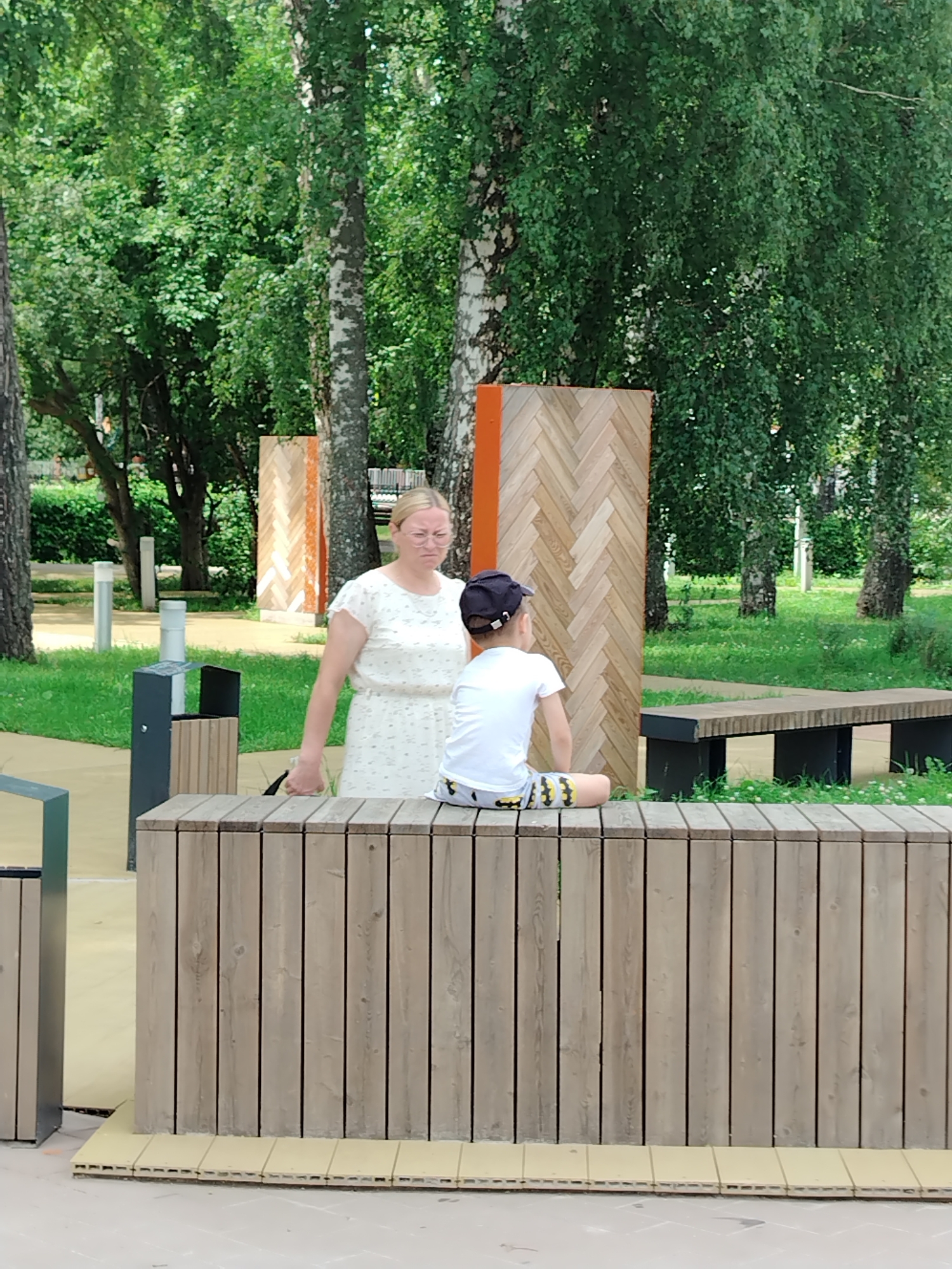 In Yanaul, Russia, a woman and a young boy are seen in an outdoor park setting. The woman, wearing glasses and a light-colored, patterned dress, stands facing away from the camera, looking towards the boy. The boy, also facing away, is seated on a wooden structure, wearing a white t-shirt and patterned shorts, with a dark baseball cap on his head. Behind them, a large, patterned wooden structure with orange accents is visible, along with trees and grassy areas. A bench is situated in the background. A trash receptacle is to the left of the woman. The scene appears to be a public space designed for leisure.