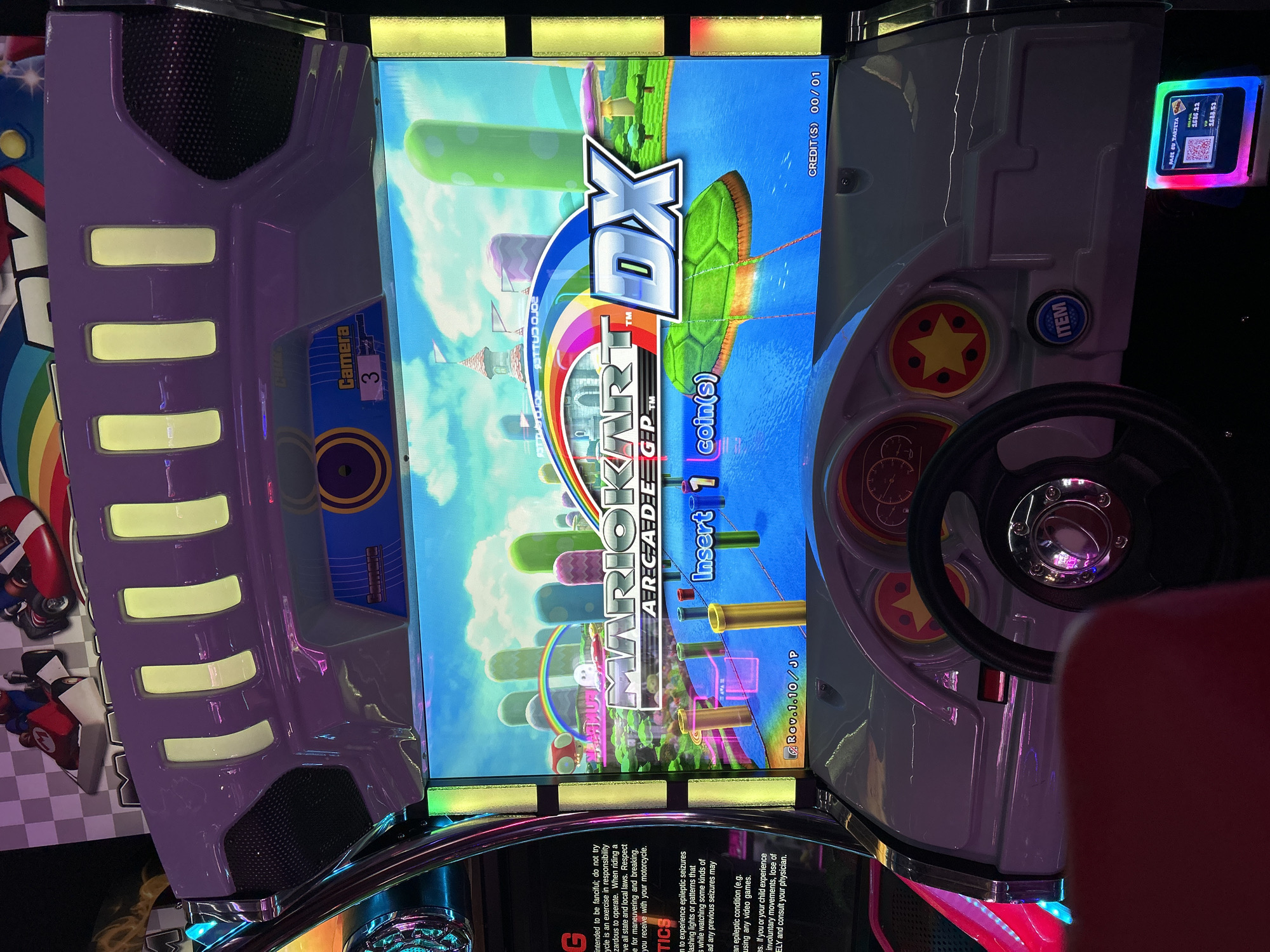An upright arcade cabinet for the game "Mario Kart Arcade GP DX" is captured in an arcade setting, likely in Buenos Aires, Argentina. The machine is in an attract mode, with its large vertical screen displaying the game's title prominently against a vibrant, cartoonish background featuring a rainbow road and a green shelled Koopa Troopa glider. The prompt "Insert 1 coin(s)" and "CREDIT(S) 00/01" indicate it's ready for gameplay.
The console is designed with a futuristic aesthetic, primarily in purple and white, accented with glowing yellow light strips. A black steering wheel is visible on the right, along with a red button featuring a yellow star and a blue "ITEM" button. A small digital display panel on the upper right shows "PAGO EU TARJETA" (Pay with Card) and numerical values "1586.22" and "1688.53", possibly related to a payment or loyalty card system, alongside a QR code. On the left side of the machine, a label reads "Camera 3". Partially visible warning text at the bottom left mentions "epileptic seizures" and advises consulting a physician. The game's version, "Rev.1.10/JP", is also displayed on the screen.
The environment is dimly lit, emphasizing the bright, colorful screen and the machine's neon accents, suggesting an indoor evening or nighttime setting typical of an arcade. No players are visible, but the setup implies an active entertainment venue awaiting engagement.