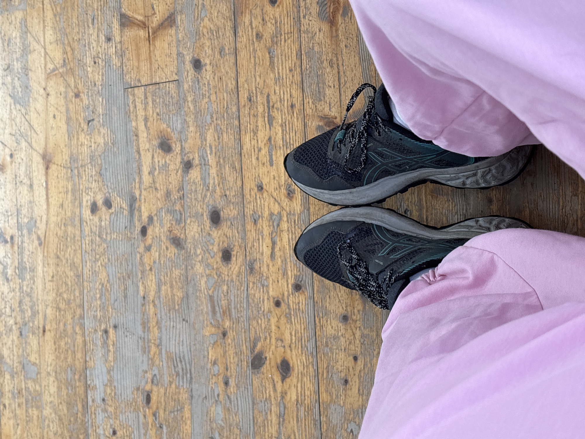 The image shows a top-down view of a person standing on a wooden floor. The person is wearing a pair of black and grey Asics running shoes and light pink pants. The wooden floor is weathered with a visible grain pattern and varying shades of brown and grey. The location cannot be determined from the image.