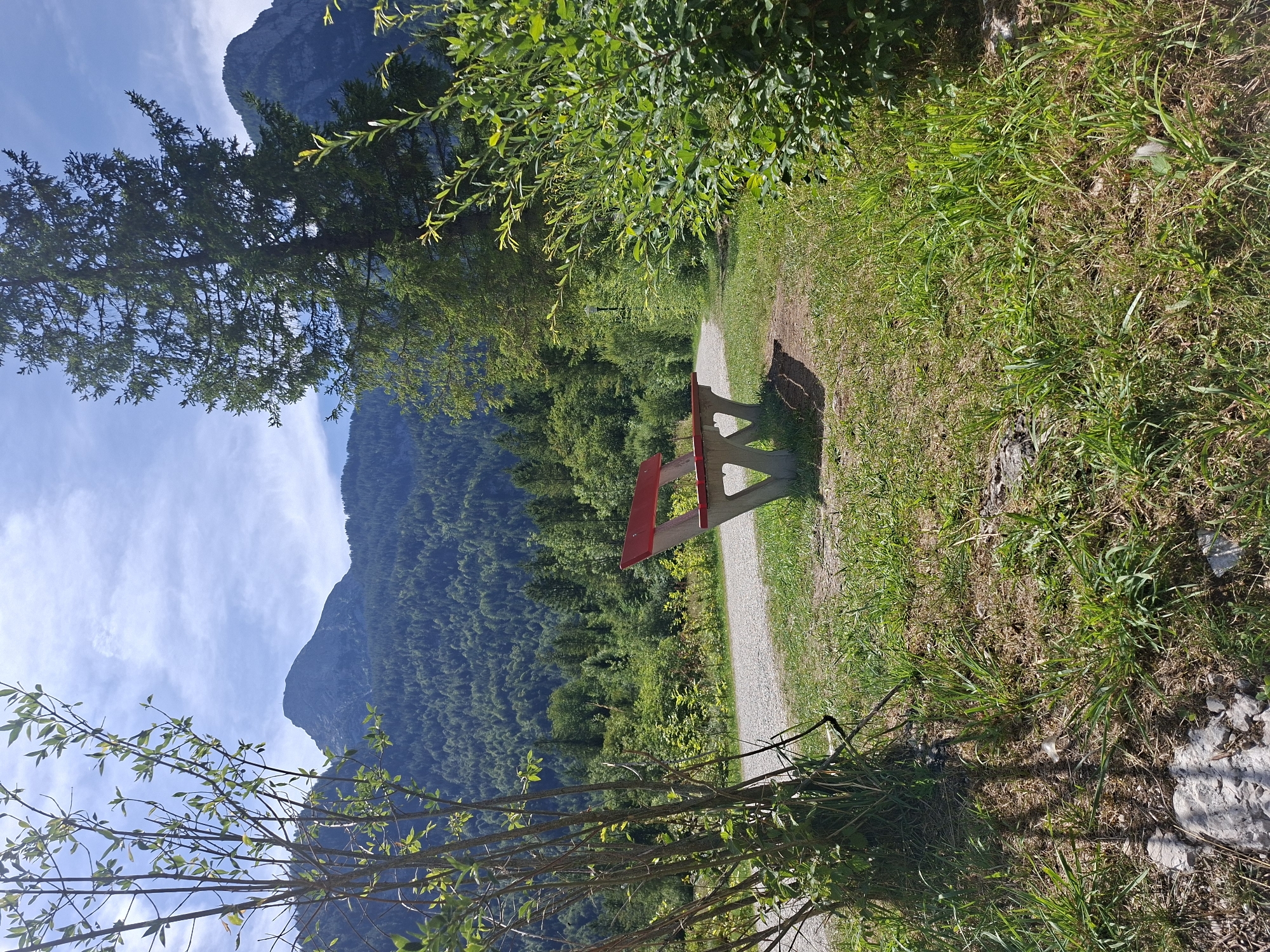A red and gray concrete bench sits on a grassy area next to a gravel path. Behind the bench, a dense forest covers a mountain slope. The sky is blue with scattered clouds. The scene appears to be in Obertraun, Austria.