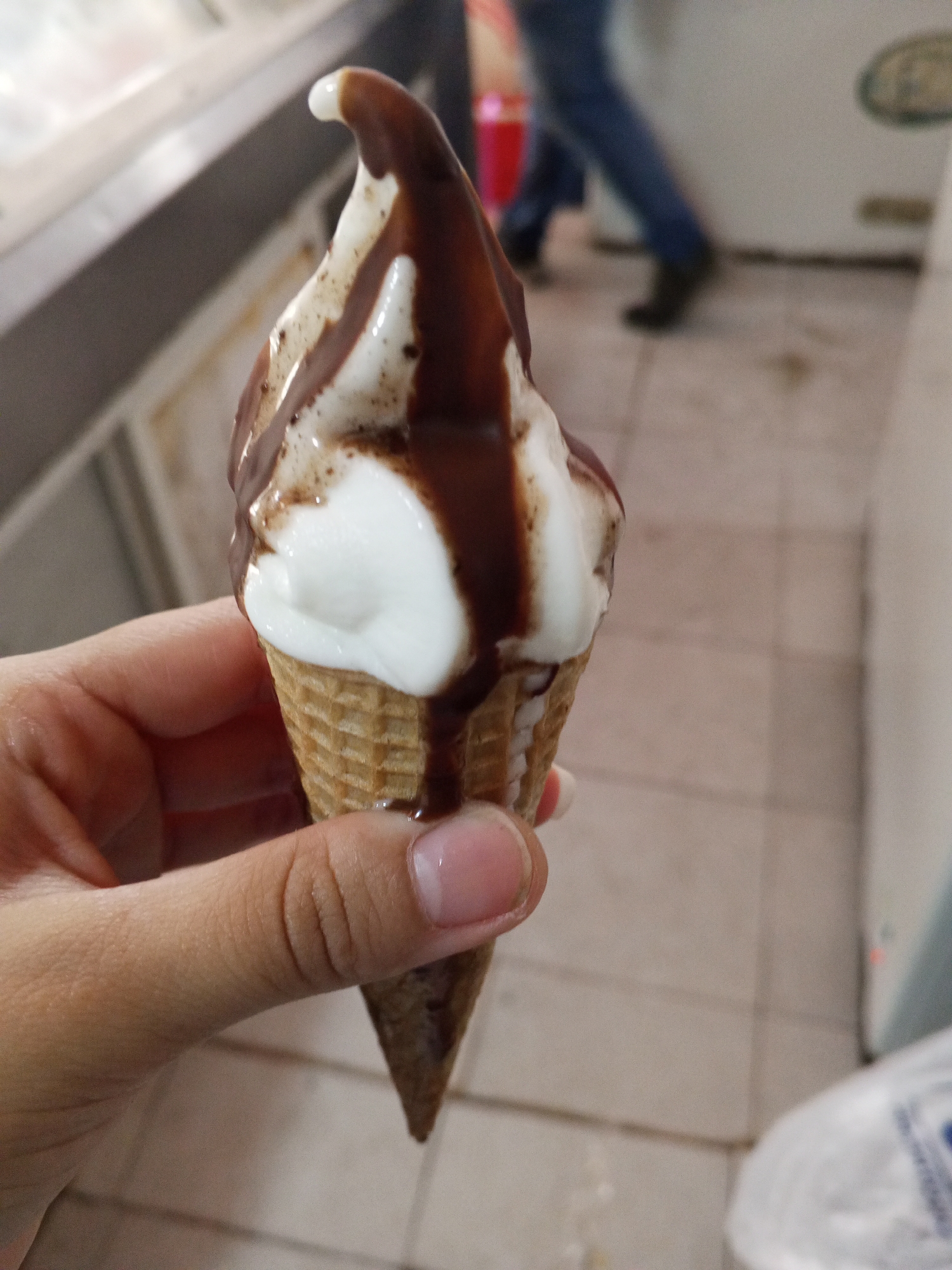 A person is holding an ice cream cone topped with chocolate sauce. The cone is a standard waffle cone filled with vanilla ice cream. The chocolate sauce is drizzled over the ice cream. The person's hand is slightly tanned and has a well-groomed nail. The background appears to be a retail establishment with tile floors and refrigeration units. There are other people faintly visible in the background. The location is Tinaquillo, Venezuela.