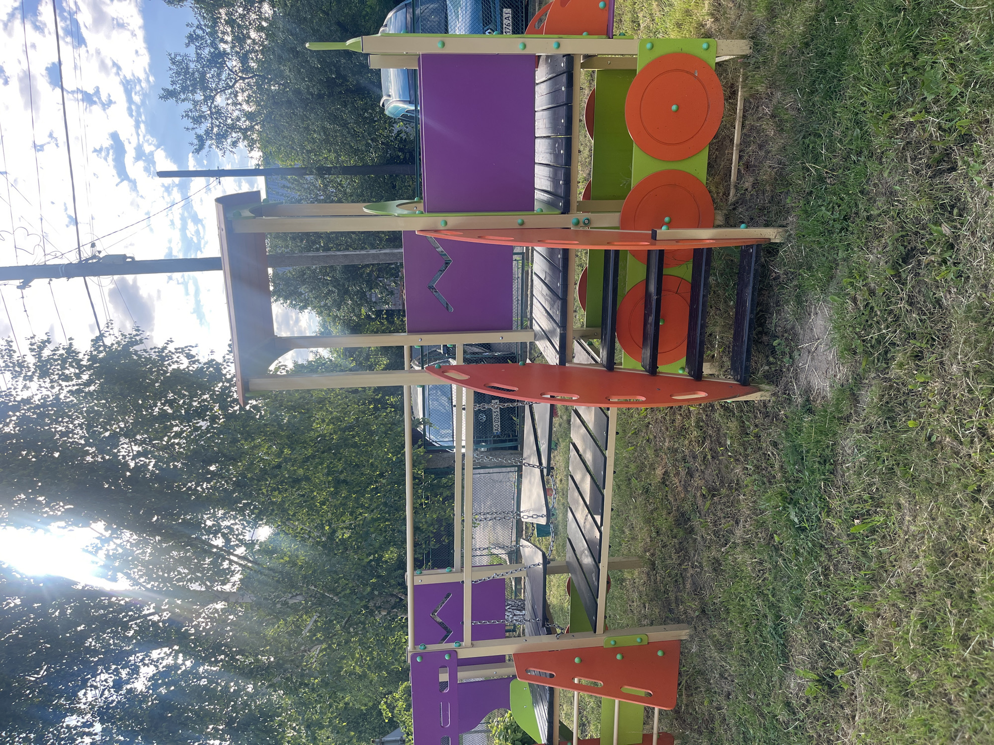The image shows a colorful playground structure resembling a train, situated on a grassy area in Botfalva, Ukraine. The playground is constructed primarily from wood and brightly colored plastic panels in purple, orange, and green. It features multiple levels, a slide, stairs, and decorative circular elements that mimic train wheels. In the background, trees and a fence are visible, along with a parked car. The sun is shining brightly, with sunbeams filtering through the leaves of a large tree in the foreground, casting light and shadows on the scene. The sky is blue with scattered clouds. There are no people or animals visible in the image.