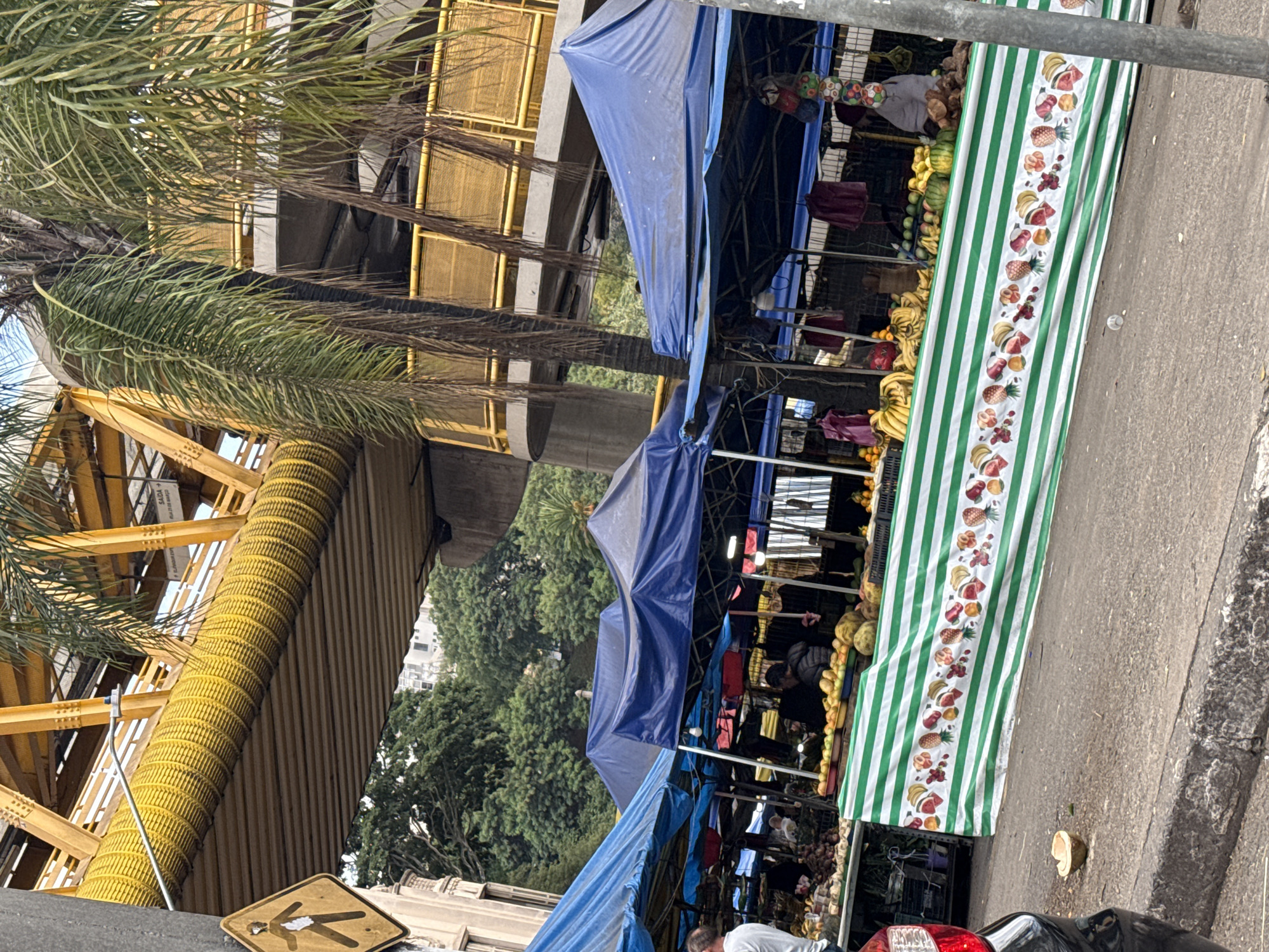 The image captures a vibrant street scene in São Paulo, Brazil, featuring a bustling outdoor fruit and vegetable market. Vendors are operating under blue tarps and striped awnings adorned with fruit patterns. An array of produce including bananas, melons, and other fruits are visibly displayed. Several people are present, presumably customers and vendors. Overhead, a pedestrian walkway with yellow corrugated piping snakes across the street, casting shadows. The background reveals lush green trees and urban buildings, typical of a Brazilian cityscape. A street sign is partially visible in the foreground. A car is parked nearby.