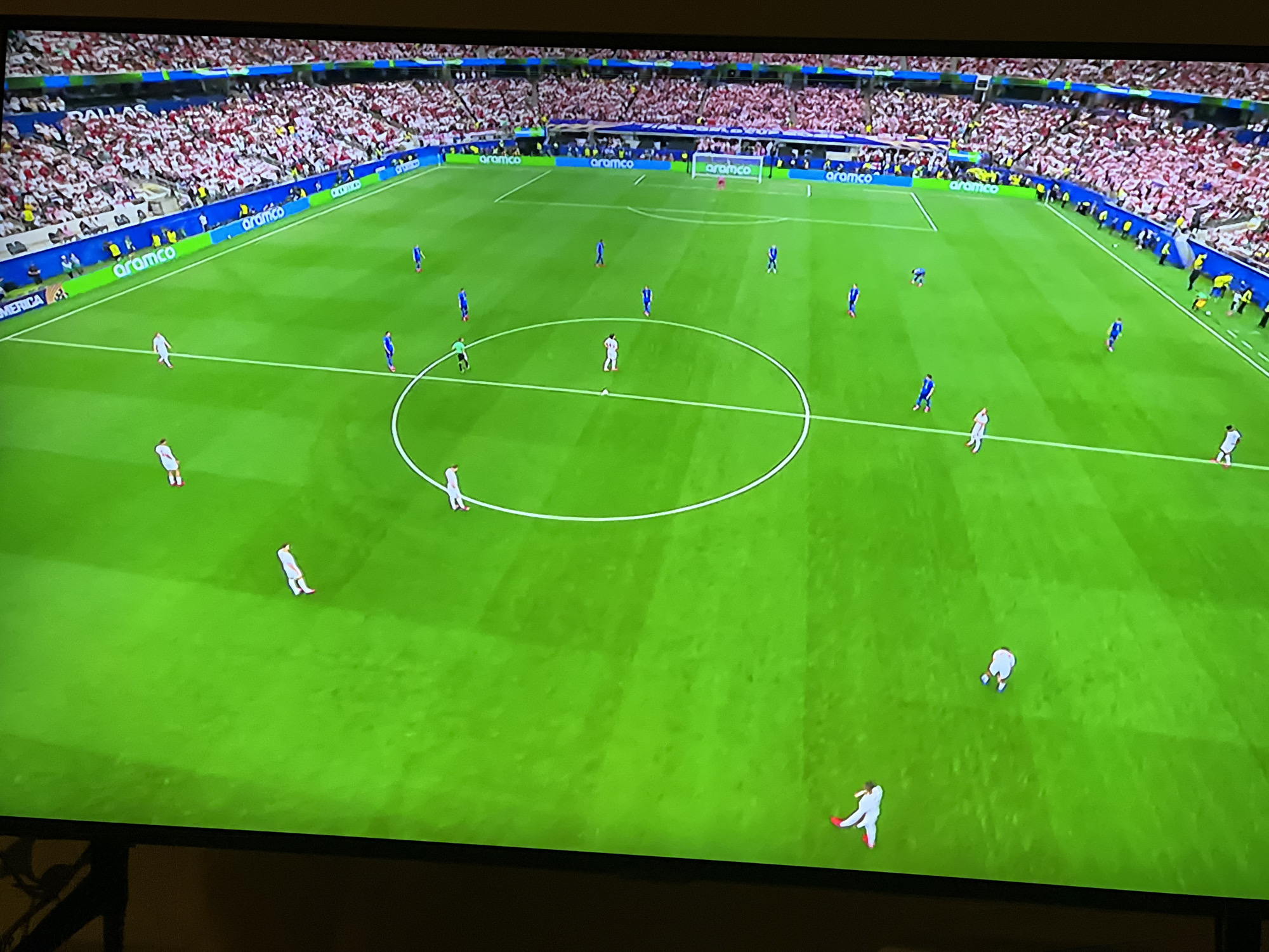 This is a high-angle, full-frame, medium shot of a professional soccer match on a brightly lit, green grass field. The field is marked with white lines, including a center circle. Spectators in red and white attire fill the stadium seating that surrounds the field. Several players, dressed in blue and white uniforms, are scattered across the field, some in motion. A large, white banner with green text that reads "aramco" runs along the perimeter of the field. The stadium appears to be modern and well-maintained. There is no indication of the city of Florești, Romania in this image.