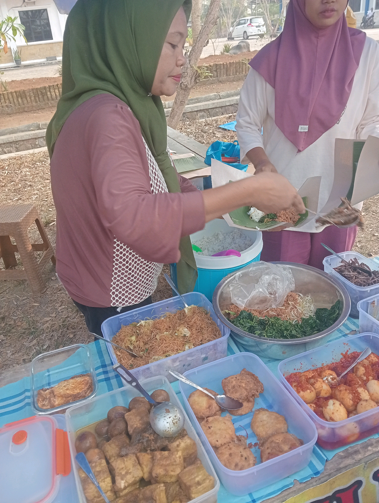 In Sidoharjo, Indonesia, a woman wearing a green hijab and a maroon shirt with a white lattice pattern is serving food from a stall. She is holding a serving spoon and a piece of wrapped food, preparing to place it onto a plate. Beside her, another person in a purple hijab and a white shirt is also handling food packaging. In front of them, a variety of dishes are displayed in plastic containers on a blue and white striped cloth. These include fried noodles, vegetables with bean sprouts and shredded meat, fried snacks, eggs in sauce, and fried tofu. The scene appears to be outdoors, with dry ground and vegetation visible in the background, along with a car and some buildings. A woven plastic stool is also present near the food stall.