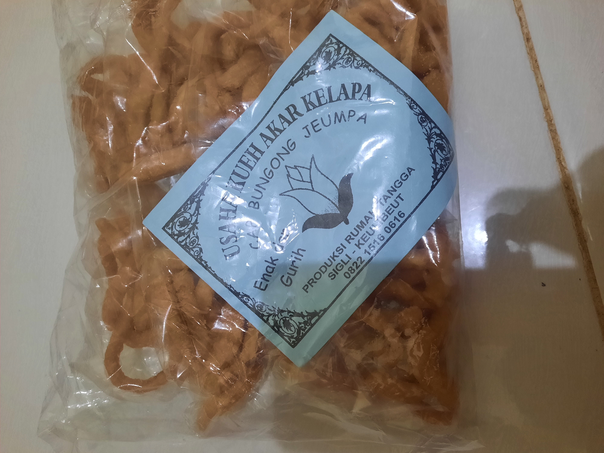 The image shows a clear plastic bag containing a large quantity of irregularly shaped, light brown fried snacks. A blue label is affixed to the front of the bag, angled to the right. The label has a decorative border and features text printed in dark blue. The text includes "USAHA KUEH AKAR KELAPA" at the top, followed by "BUNGONG J'EUMPA" and then "Enak Gurih". Below this, there is a small graphic of a stylized bird or flower. Further down, the label reads "PRODUKSI RUMAH TANGGA" and then "SIGLI KEU BEUT" and a phone number "0822 1516 0616". The snacks appear to be crispy and have a twisted or knotted appearance. The background is a neutral, off-white surface, possibly a table or floor, with a faint seam running horizontally across the bottom. The lighting suggests an indoor environment with overhead illumination. The location of Lhokseumawe, Indonesia is inferred from the text on the label which includes "J'eumpa", a type of flower native to Aceh, the province where Lhokseumawe is located.
