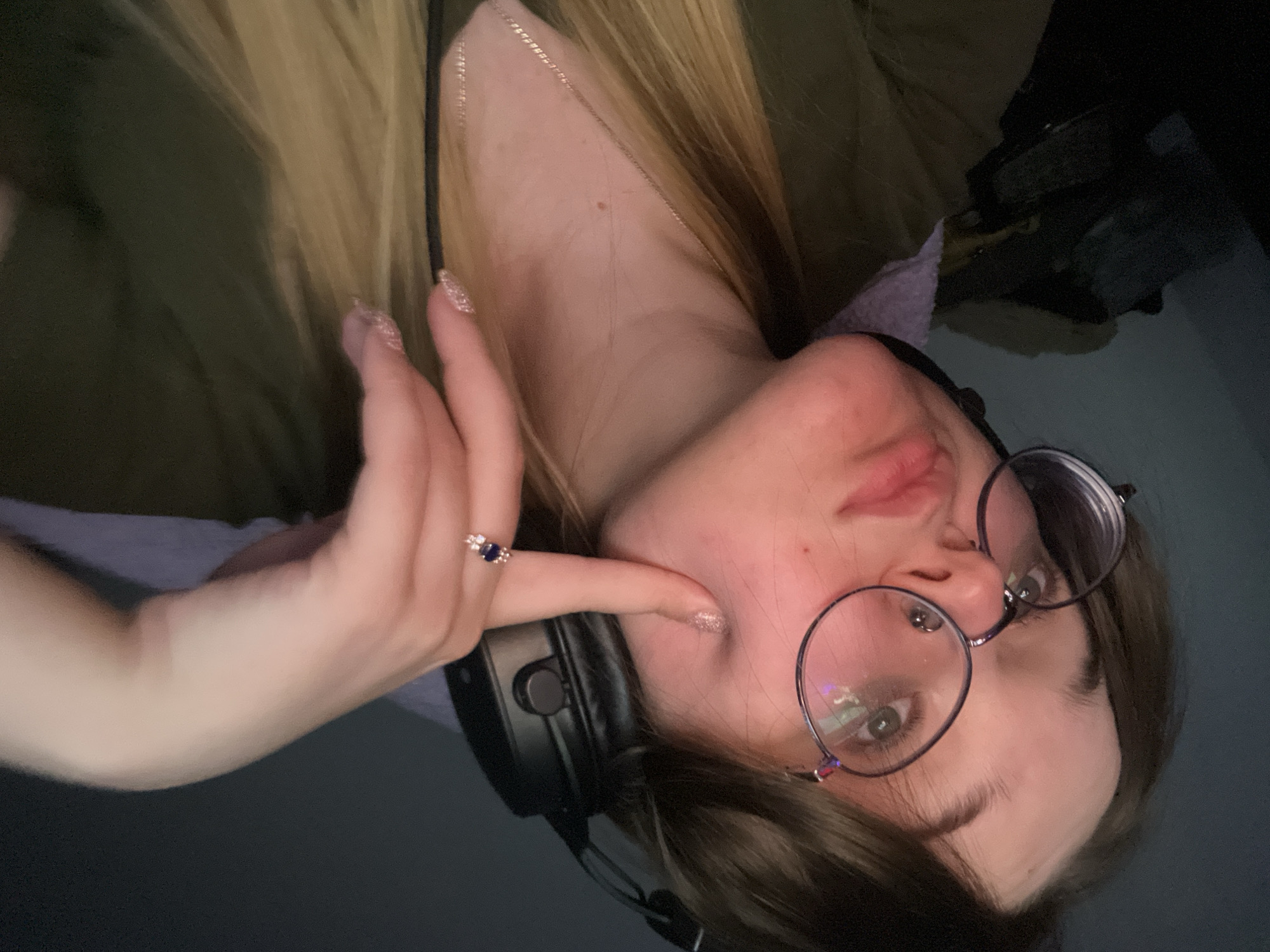 The image depicts a female individual in an indoor setting. The specific location in Pushchino, Russia, cannot be determined from the visual information provided. The subject has long, light-colored hair and wears round spectacles with dark frames and a septum piercing. Black over-ear headphones are worn. She is clad in an olive green upper garment, possibly over a light purple layer, and a thin silver-colored necklace. Her left hand is raised, with the index finger pressing into her left cheek, creating a distinct indentation. A ring with a dark blue oval stone flanked by smaller clear stones is visible on her left ring finger. Her fingernails are manicured with a glitter-detailed polish. The background is dark and out of focus, showing indistinct shapes in the lower left portion of the frame. The subject looks directly towards the viewer, illuminated by a frontal light source.