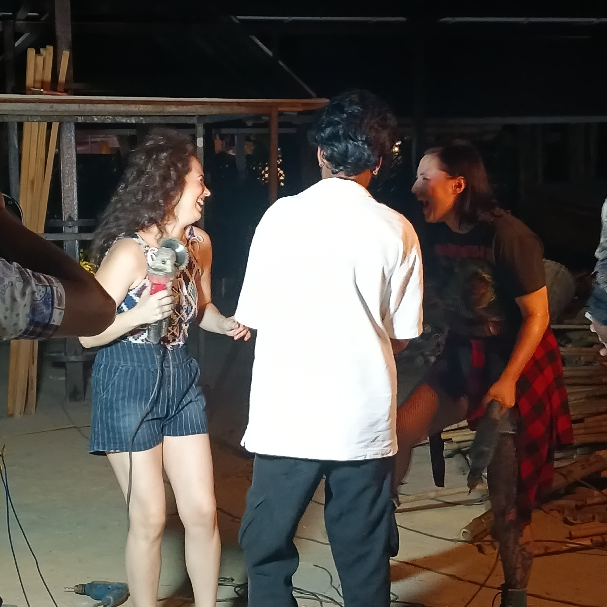 In a dimly lit indoor space in Panaji, India, two women flank a man. The woman on the left holds an angle grinder and laughs, while the woman on the right, wearing a t-shirt and plaid shirt, gestures emphatically. The man stands between them, his back to the viewer, wearing a white shirt and dark pants. A drill rests on the floor. Stacks of lumber are in the background.