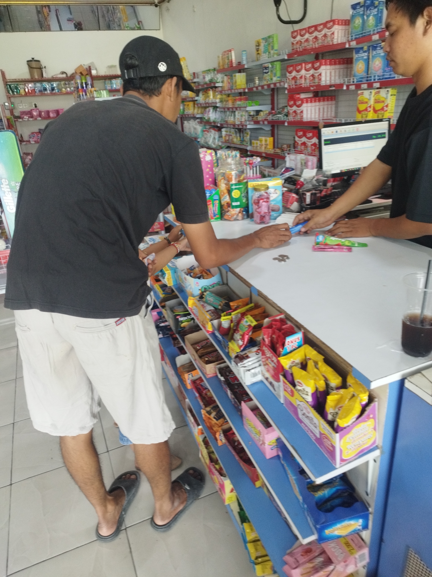 The image depicts the interior of a small convenience store, likely a warung, in Woro, Indonesia. A man wearing a black t-shirt and light-colored shorts stands behind a counter, facing away from the camera. He is wearing a black baseball cap. Another person, partially visible, is on the other side of the counter, appearing to interact with the first man. The counter itself is white and displays various items, including what appears to be money and a blue pen. Shelves are visible behind the counter, stocked with an assortment of packaged goods, including snacks and drinks. A computer monitor is also present on the counter. The flooring is tiled with a light-colored pattern. The overall impression is that of a typical retail transaction taking place in a local Indonesian shop.