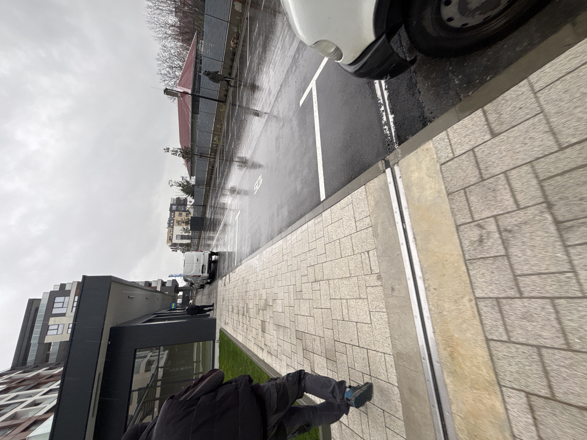 The image shows an outdoor scene on a rainy day, possibly in Plovdiv, Bulgaria, though this is not explicitly confirmed by visual elements. The primary view is of a wet parking lot with white parking lines. A white van partially obscures the upper right corner of the frame. In the distance, other parked vehicles are visible, along with a person walking in the parking area.
A sidewalk paved with rectangular stones runs along the bottom and right sides of the frame. A person is partially visible at the bottom of the image, wearing dark clothing and walking on the sidewalk. Adjacent to the sidewalk, there is a narrow strip of green grass.
A multi-story building with modern architecture is visible on the left side of the frame. Another building with a red roof and metal fencing is located in the background on the left. The sky is overcast.