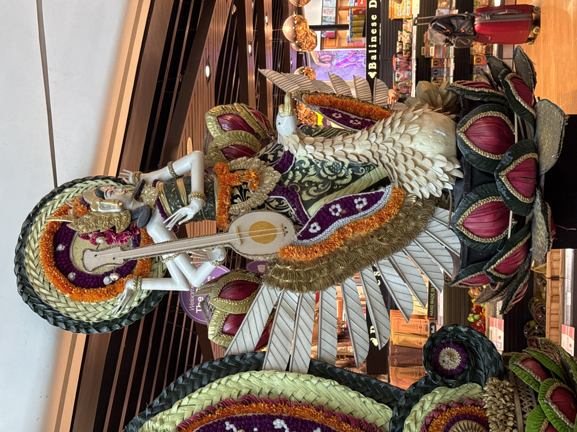 The image shows a large, ornate statue depicting a deity with multiple arms playing a stringed instrument, seated on a large swan. The statue is adorned with colorful floral and woven decorations. It is situated in an indoor environment, likely an airport or a large retail space, with shelves of products visible in the background. The overall scene suggests a cultural display, possibly in Kuta, Indonesia, a popular tourist destination known for its vibrant arts and crafts. The statue's elaborate design and the surrounding environment indicate a place of commerce and cultural exhibition, where travelers and shoppers might encounter such artistic representations.