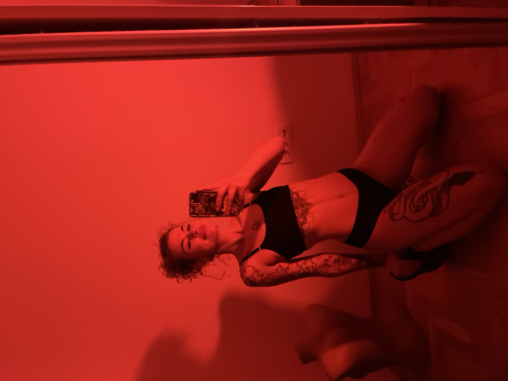 A person is visible in a selfie taken in a room with red lighting. The person is wearing a black bra and black underwear. The person has tattoos on their torso and left arm, and curly hair. The person is holding a phone, appearing to take the selfie. There is a wall behind the person, featuring an electrical outlet. The floor appears to be tiled. The location is likely indoors, possibly in a private residence. No elements indicate this image was taken in Boreti, Montenegro.